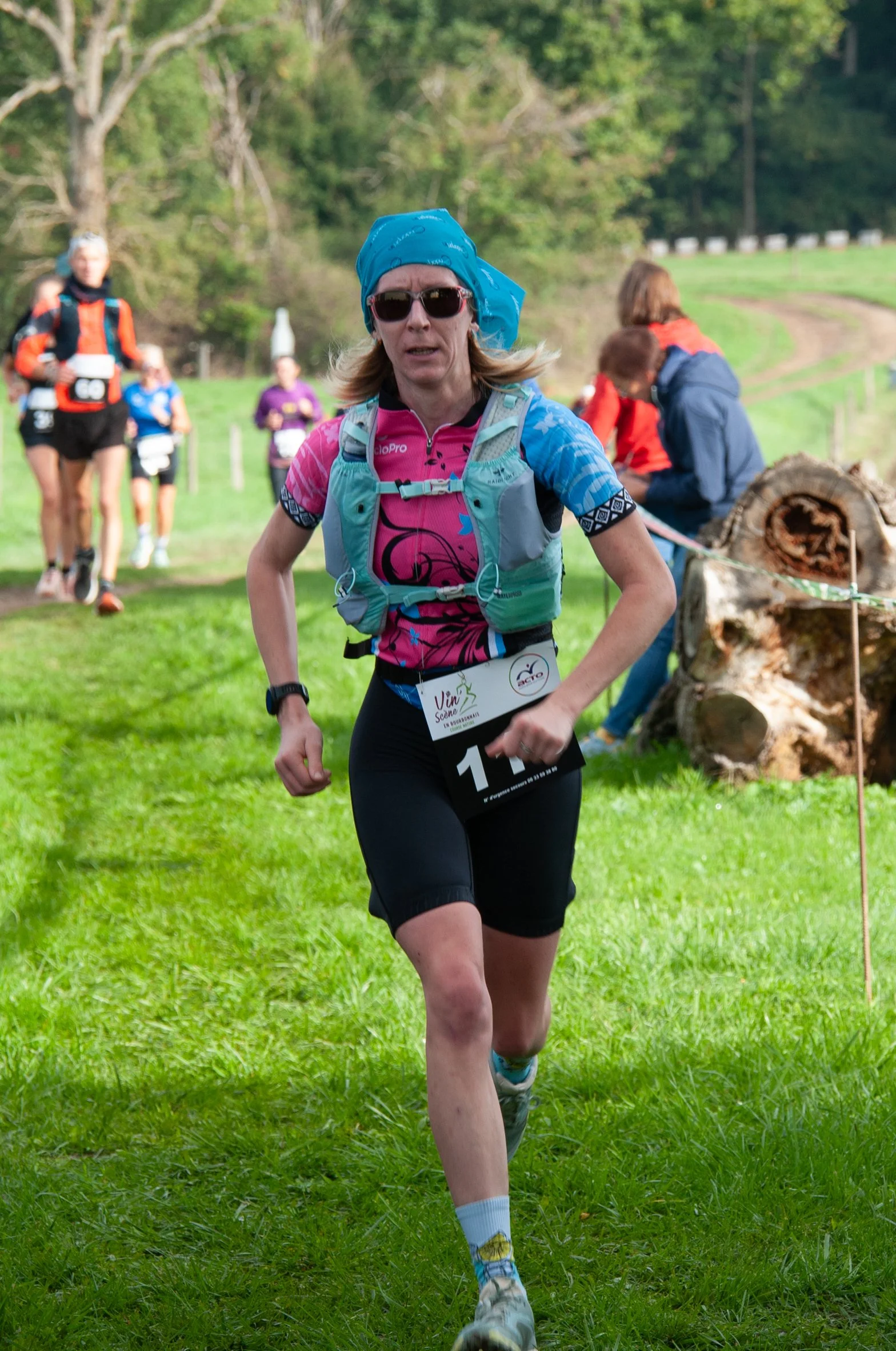 Femme courant lors d'une course dans un environnement naturel avec arbres et herbe, portant un t-shirt coloré, un bandeau bleu, des lunettes de soleil, un sac à dos et une plaque de course. D'autres participants en course sont visibles en arrière-pla