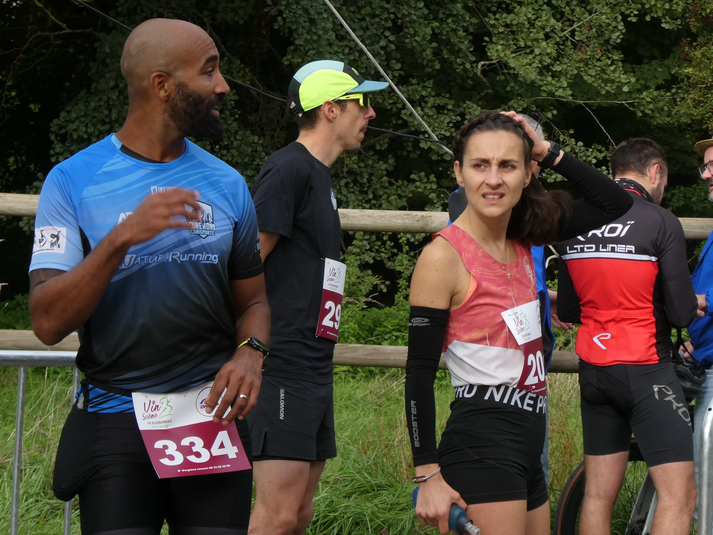 Groupe de coureurs décontractés lors d'une course en plein air avec des numéros de course, forestière en arrière-plan, un homme noir avec barbe, une femme avec un t-shirt enroulé et une autre personne en arrière-plan réfléchie.