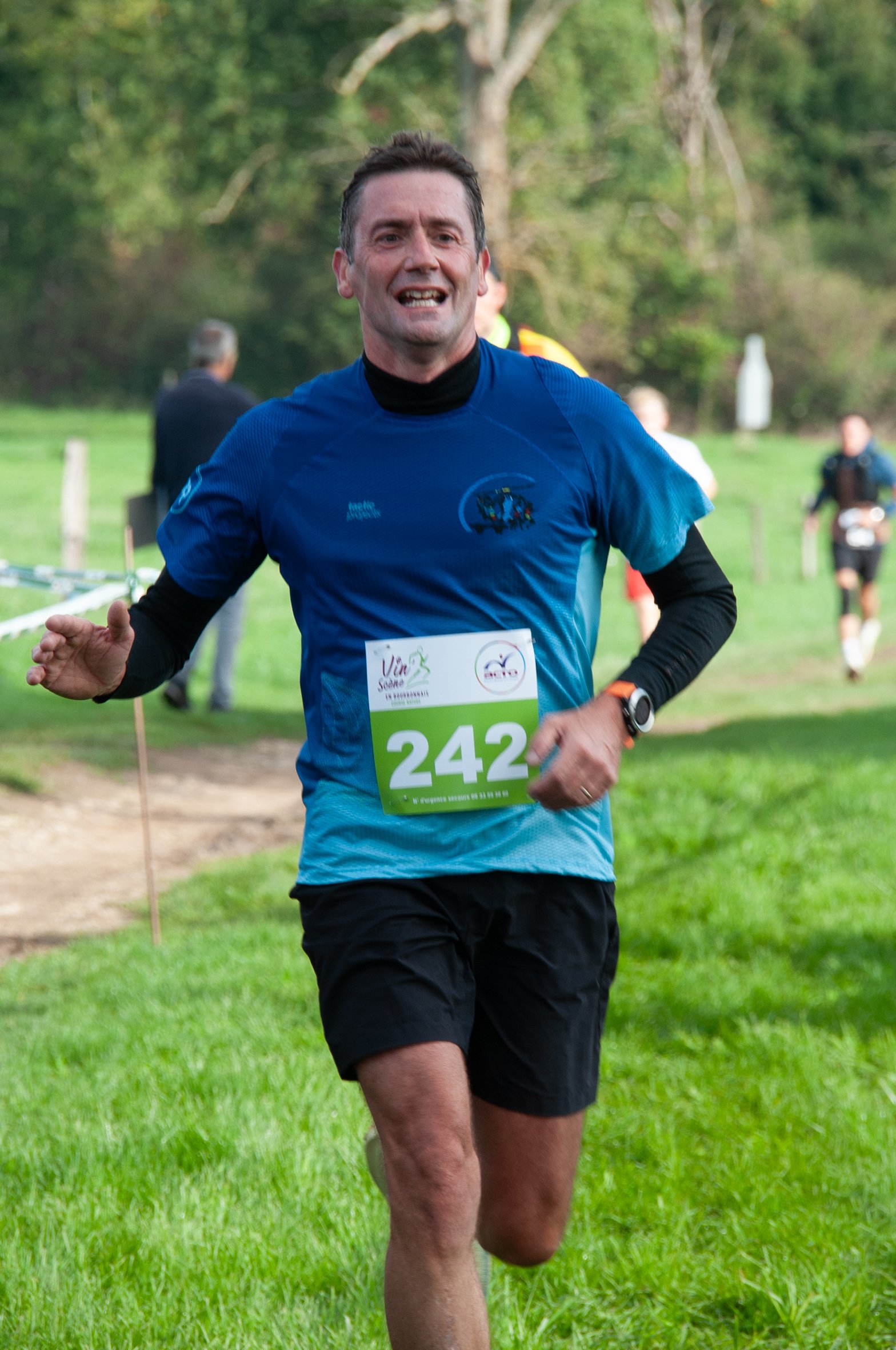 Un homme courant lors d'une course en plein air, portant un t-shirt bleu, un numéro de course 242, et une montre connectée, dans un environnement verdoyant avec d'autres coureurs en arrière-plan.