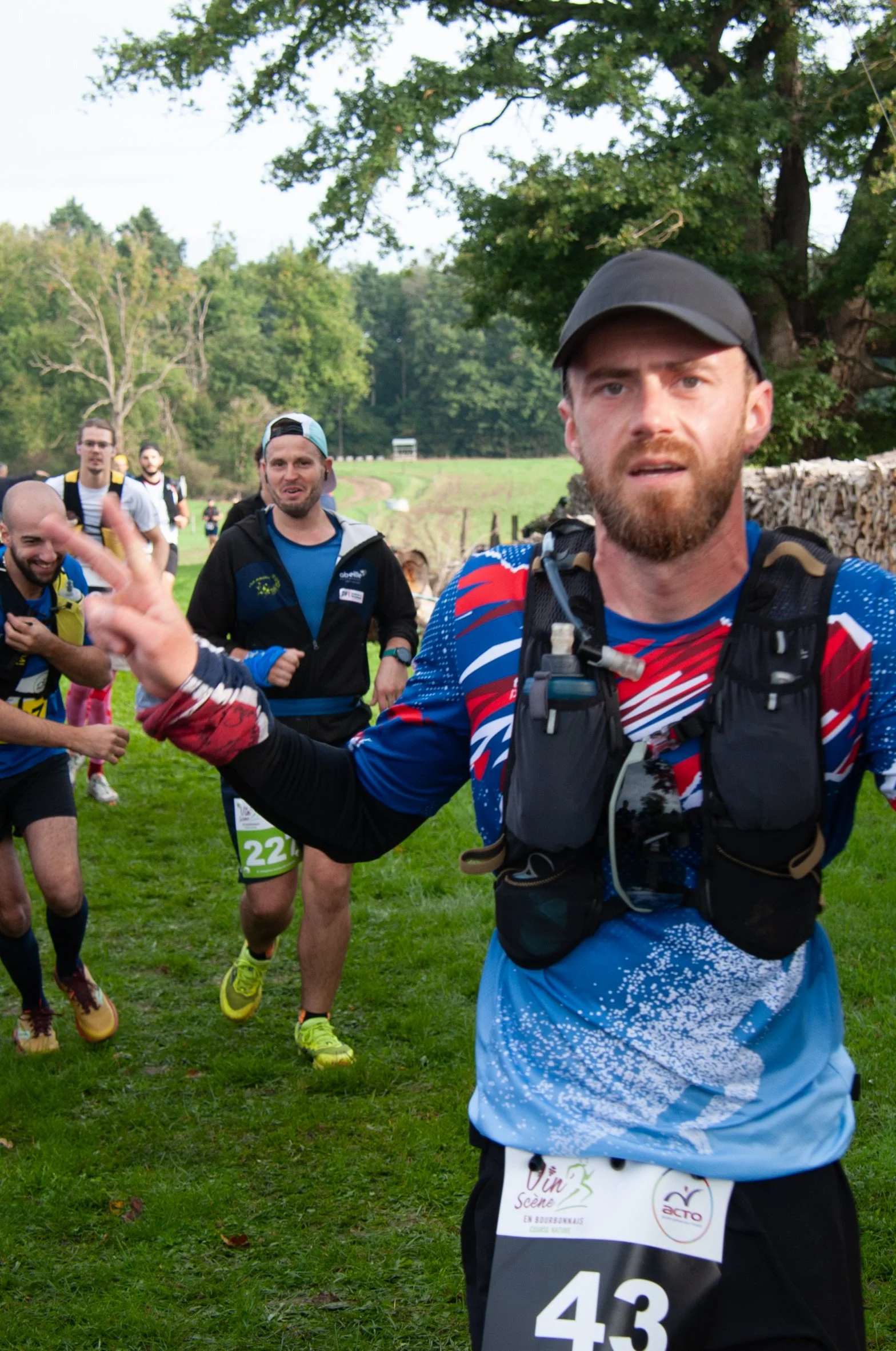 Groupe de coureurs lors d'une course en extérieur, avec un homme au premier plan portant un gilet de course, lançant un signe de paix, dans un environnement naturel avec des arbres et un ciel clair.