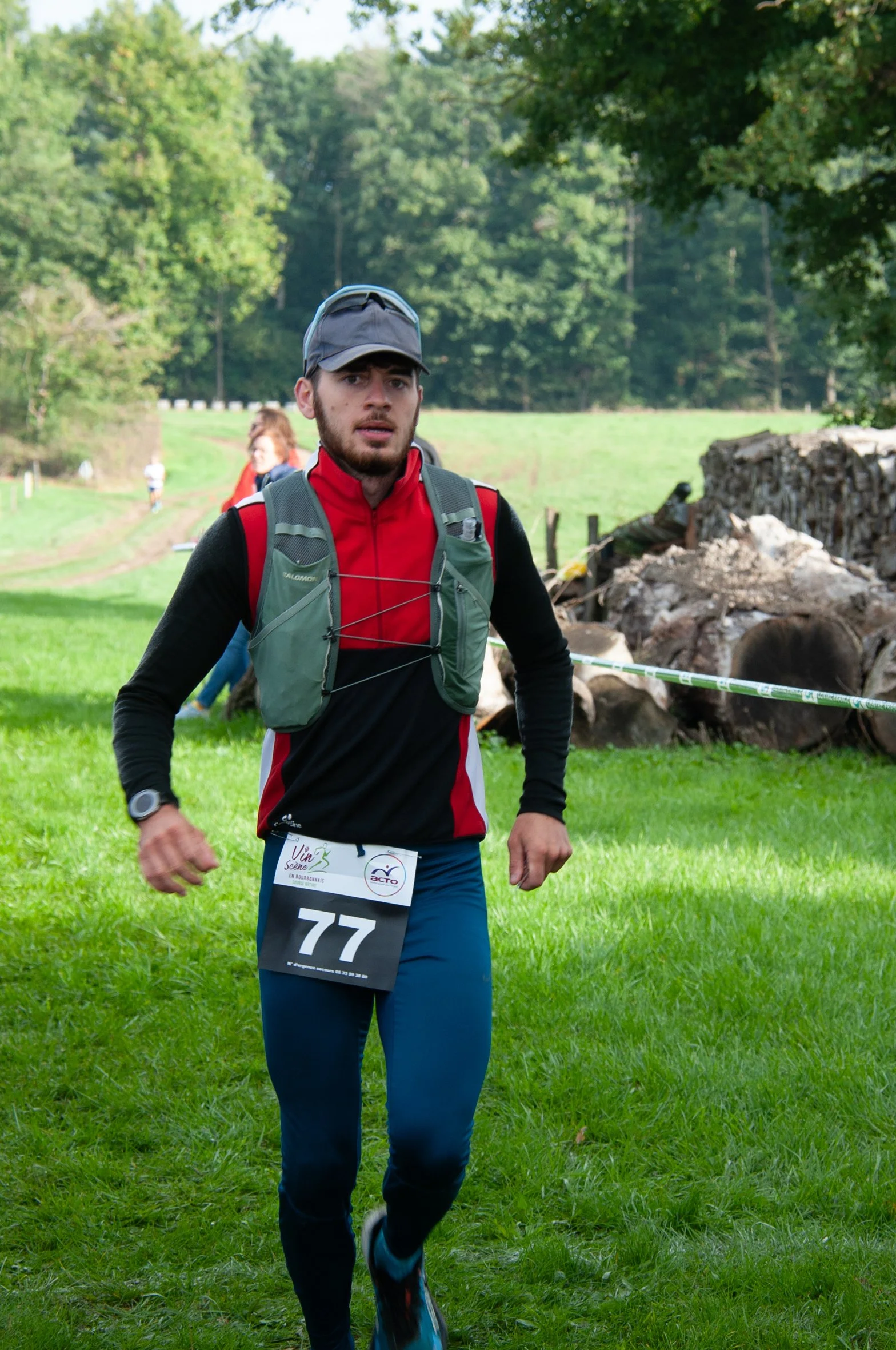 Un homme court dans un champ lors d'une course en pleine nature, portant un maillot noir et rouge, un sac à dos gris, une casquette et une montre de course, avec des autres participants en arrière-plan.