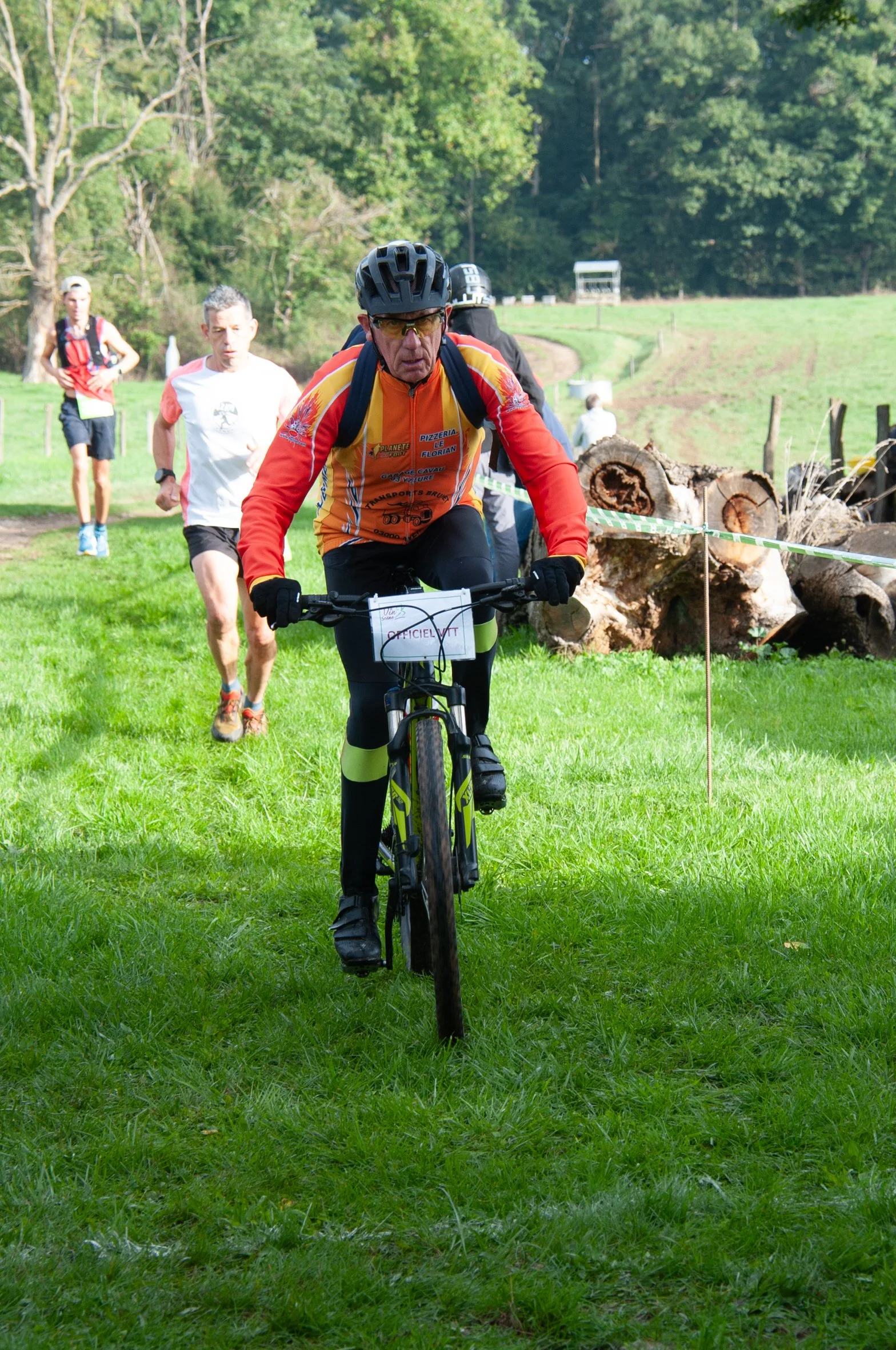 Un homme en vélo tout-terrain portant un casque, une veste orange et noire, participe à un événement en plein air dans un espace vert, avec d'autres personnes en arrière-plan.