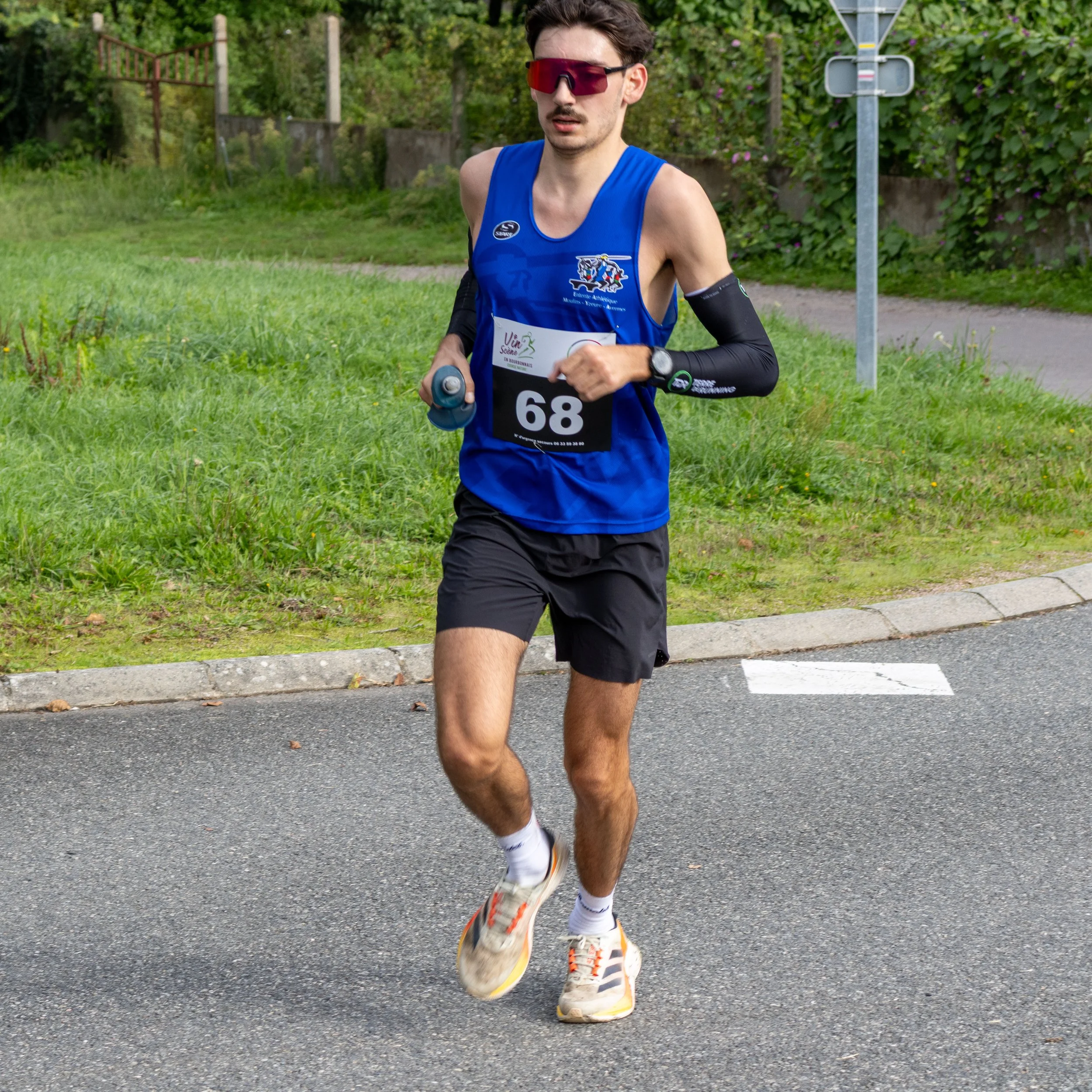 Un homme court en portant un numéro 68 lors d'une course. Il porte des lunettes de soleil, un haut sans manches bleu, des shorts noirs, des chaussures de course, et une montre.