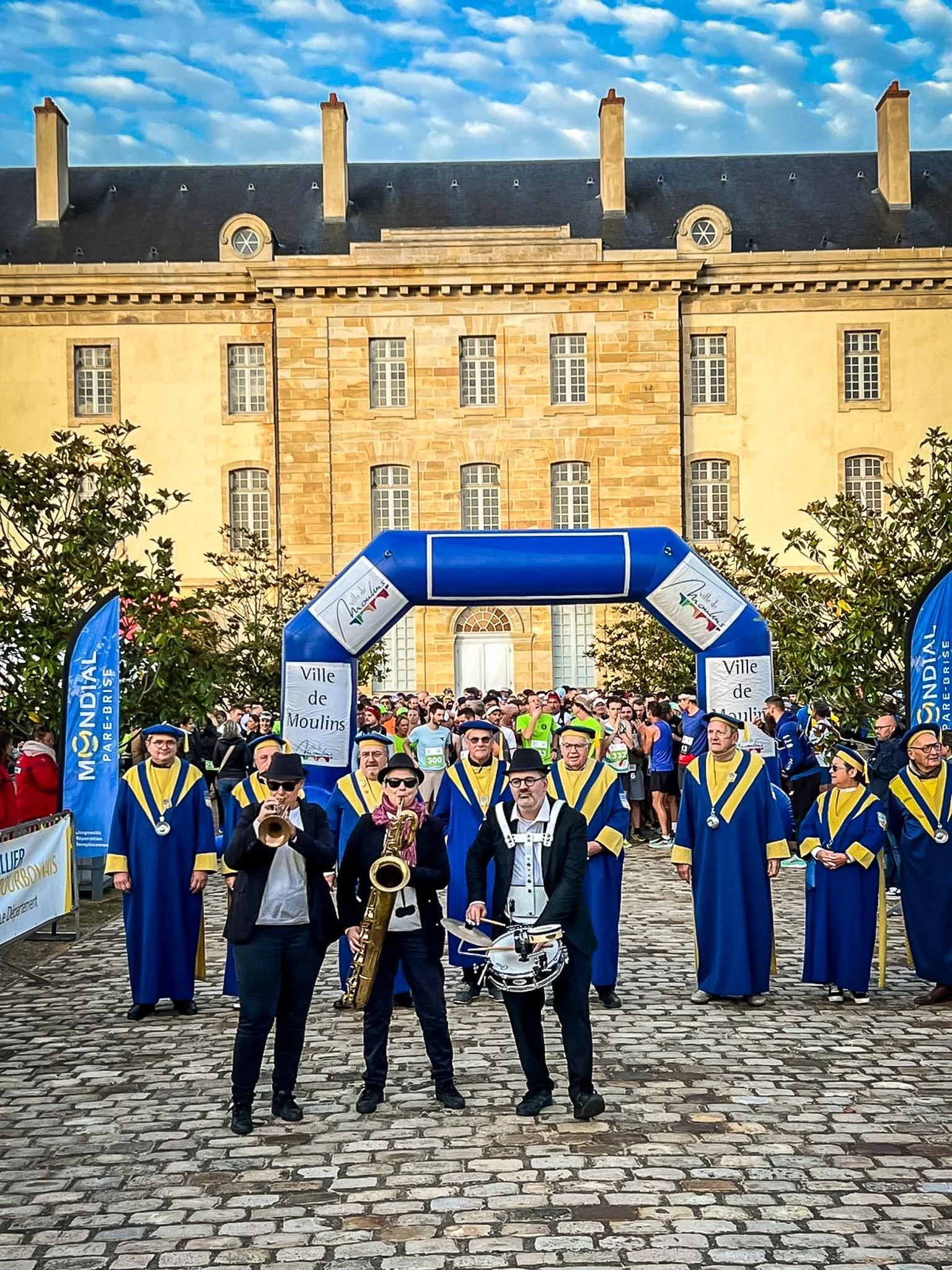 Une procession ou un événement festif dans une ville avec une bande musicale en tenue bleue et jaune, sous un arc gonflable avec le texte "Ville de Moulins". Il y a beaucoup de personnes, des bâtiments historiques en arrière-plan, et le ciel est partiellement nuageux.