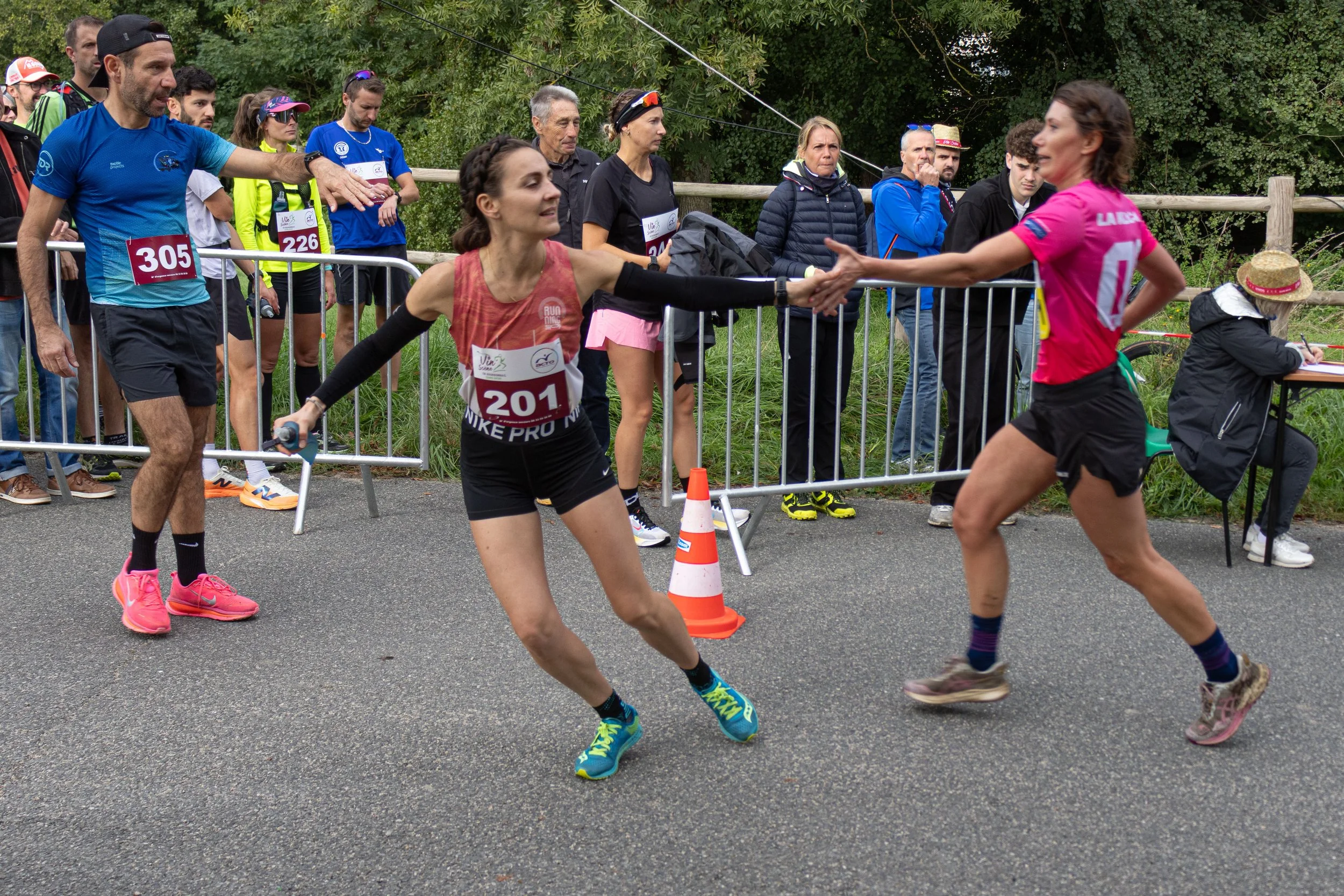 Deux femmes se font un handshake en courant lors d'une compétition de course, avec un groupe de spectateurs derrière une barrière.