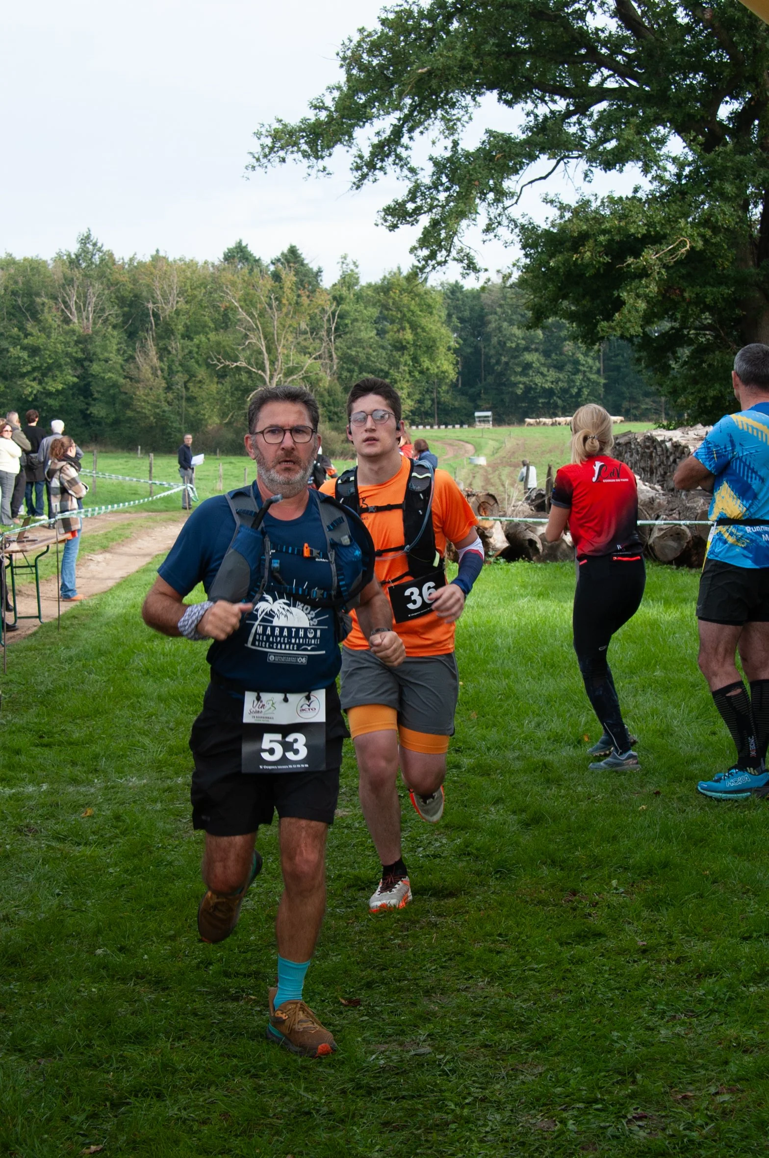 Deux coureurs participant à une course en nature, portant des numéros 53 et 36, entourés de spectateurs et de la nature verdoyante.