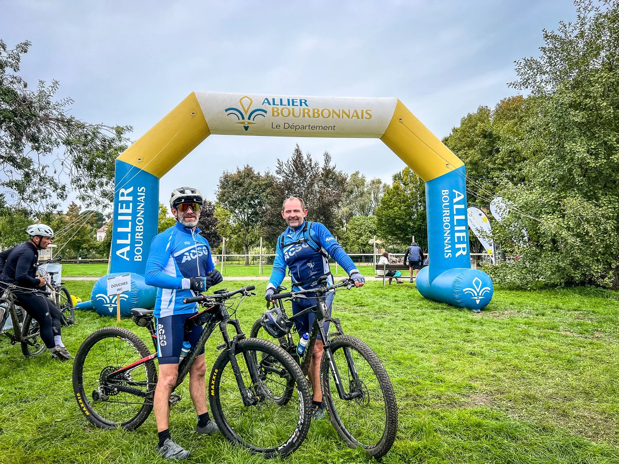 Deux hommes en tenue de cyclisme posent avec leurs vélos sous une arche gonflable indiquant Allier Bourbonnais, dans un parc, avec d'autres cyclistes en arrière-plan.