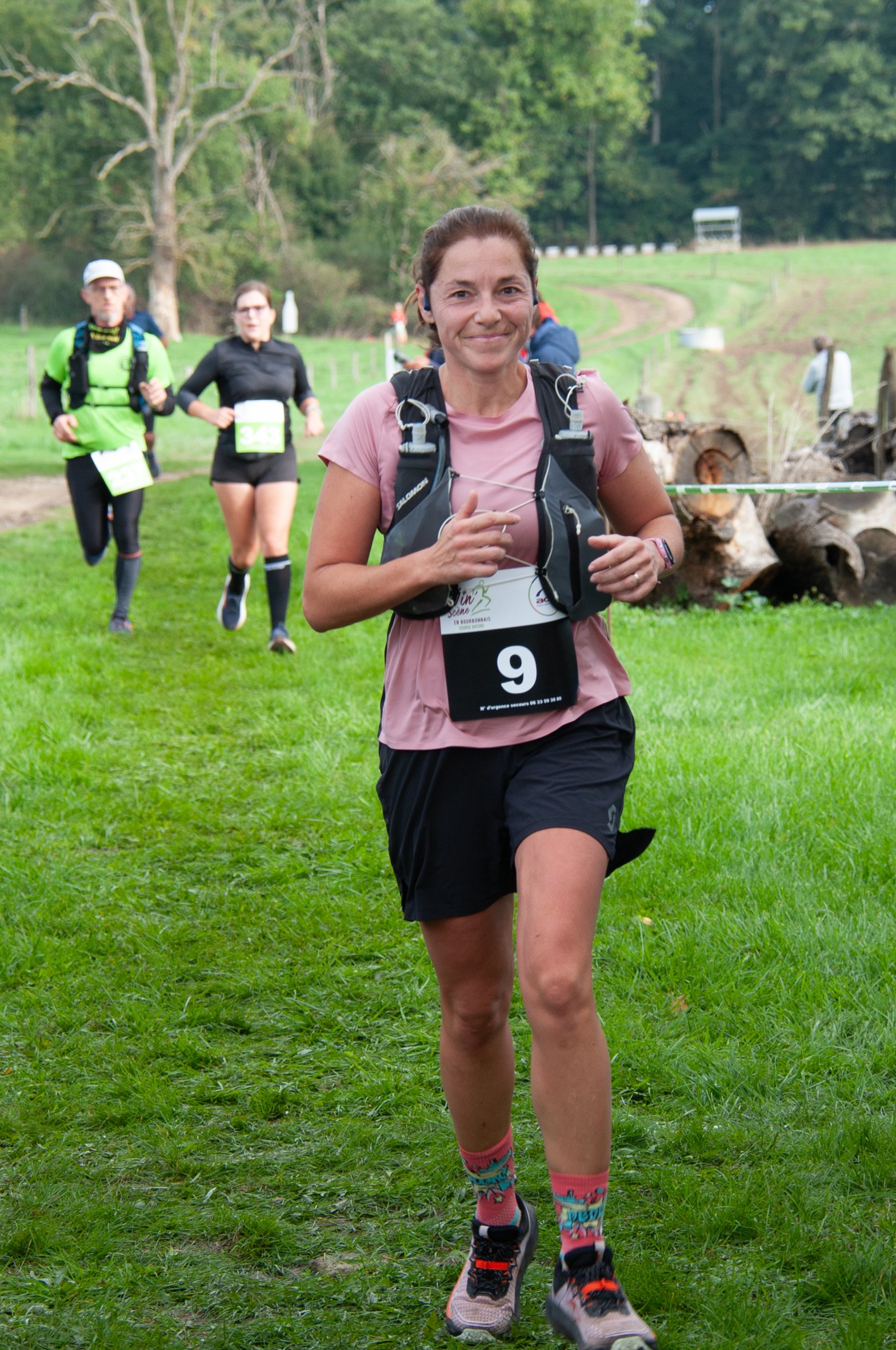 Femme courant lors d'une course en plein air, portant un dossard avec le numéro 9, dans un environnement verdoyant avec d'autres coureurs en arrière-plan.