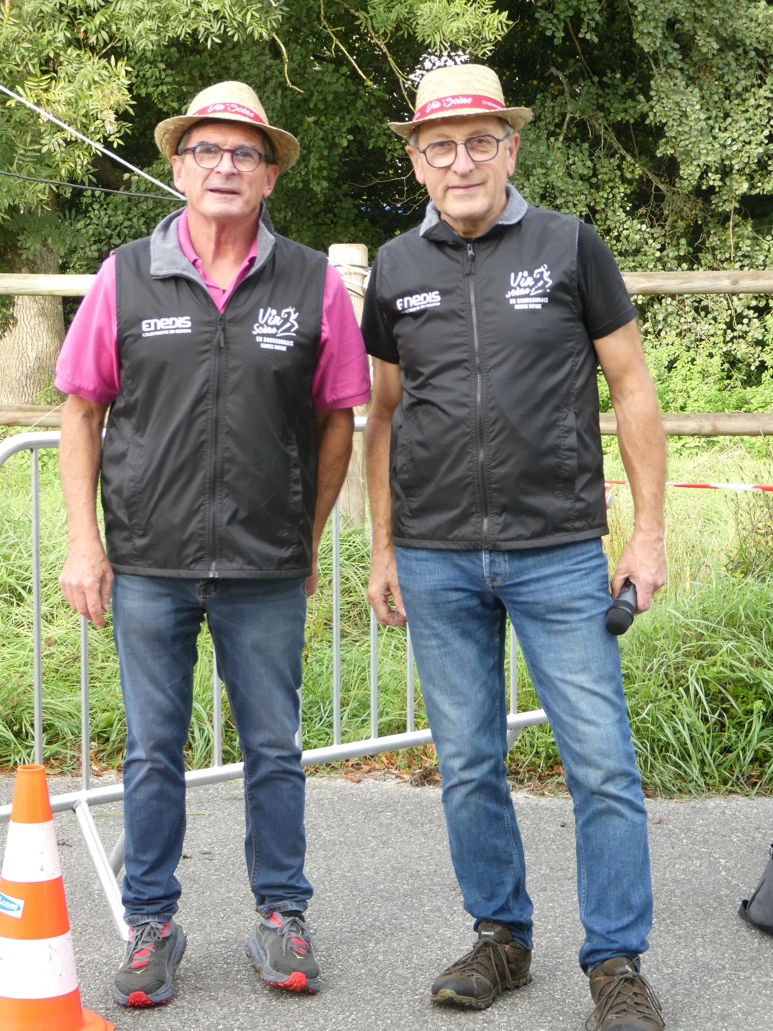 Deux hommes portant des chapeaux de paille et des gilets noirs avec des logos, debout devant une barrière en extérieur, l'un d'eux tenant un micro.
