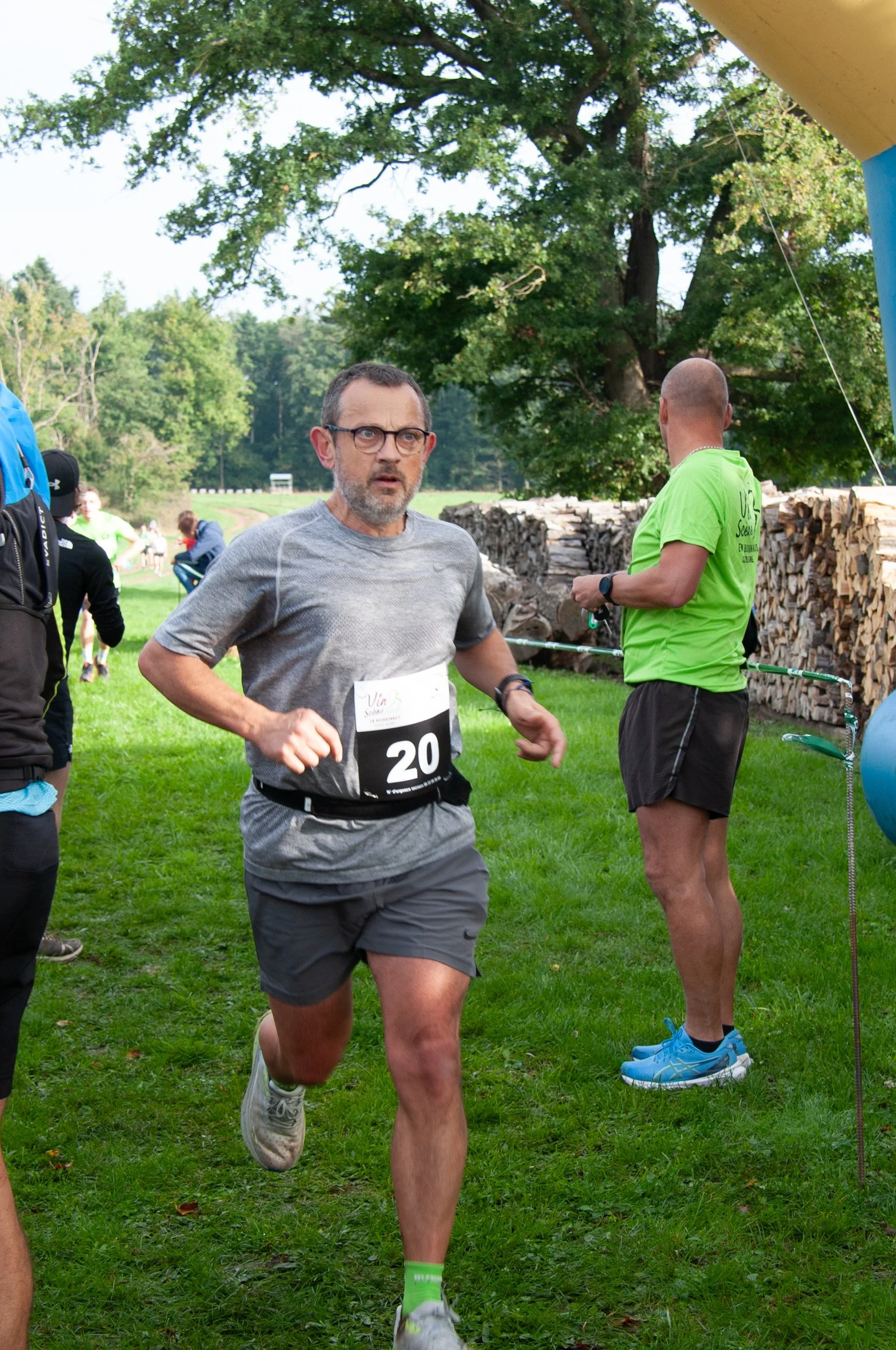 Un homme court lors d'une course en plein air, portant un T-shirt gris, un short gris, des chaussures de course, et un numéro de course 20. Il y a d'autres personnes en arrière-plan, dont un homme en T-shirt vert, qui attend ou se prépare à courir, d
