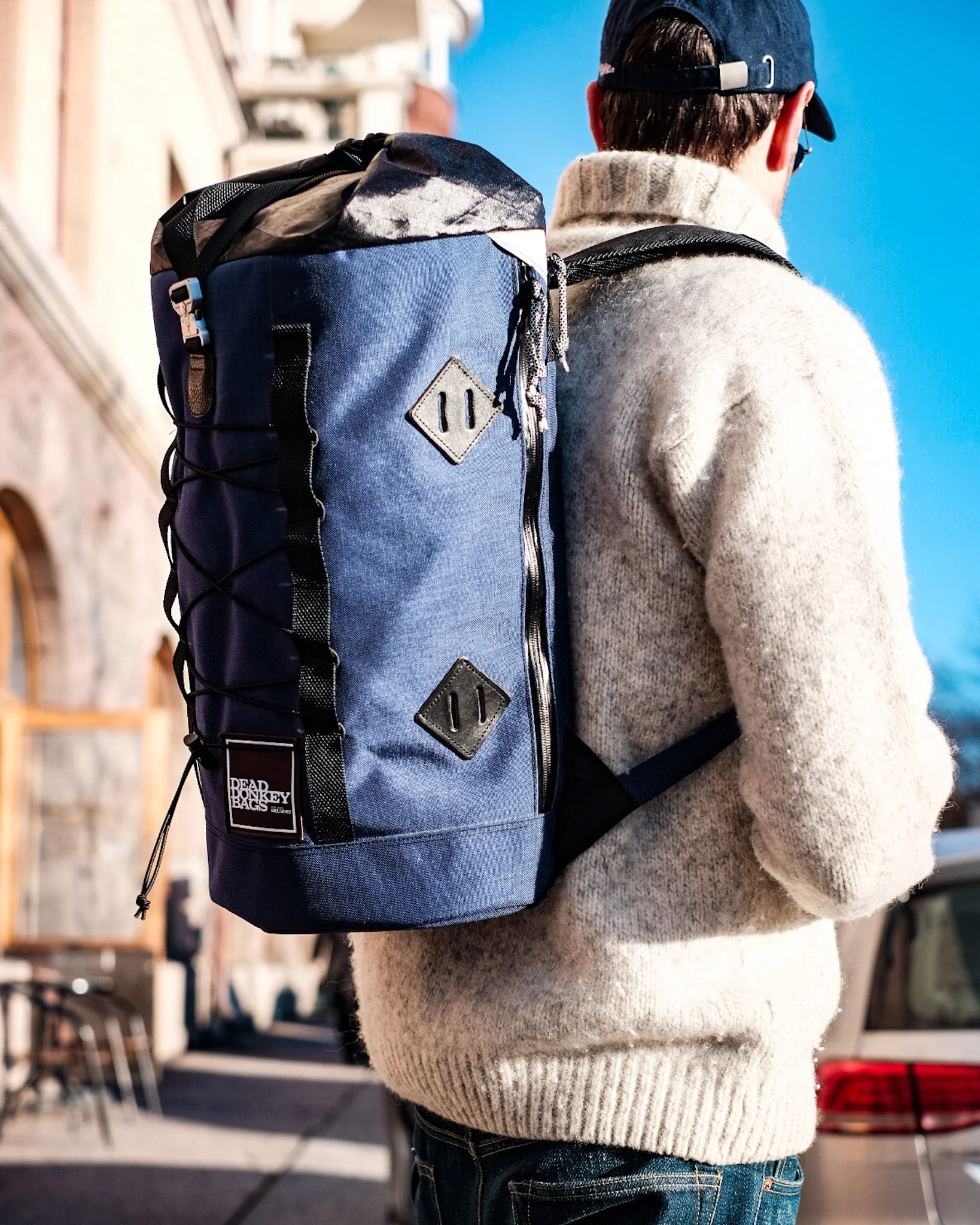 Person wearing a gray sweater, baseball cap, and sunglasses with a large blue backpack on their back, walking outdoors on a sunny day.