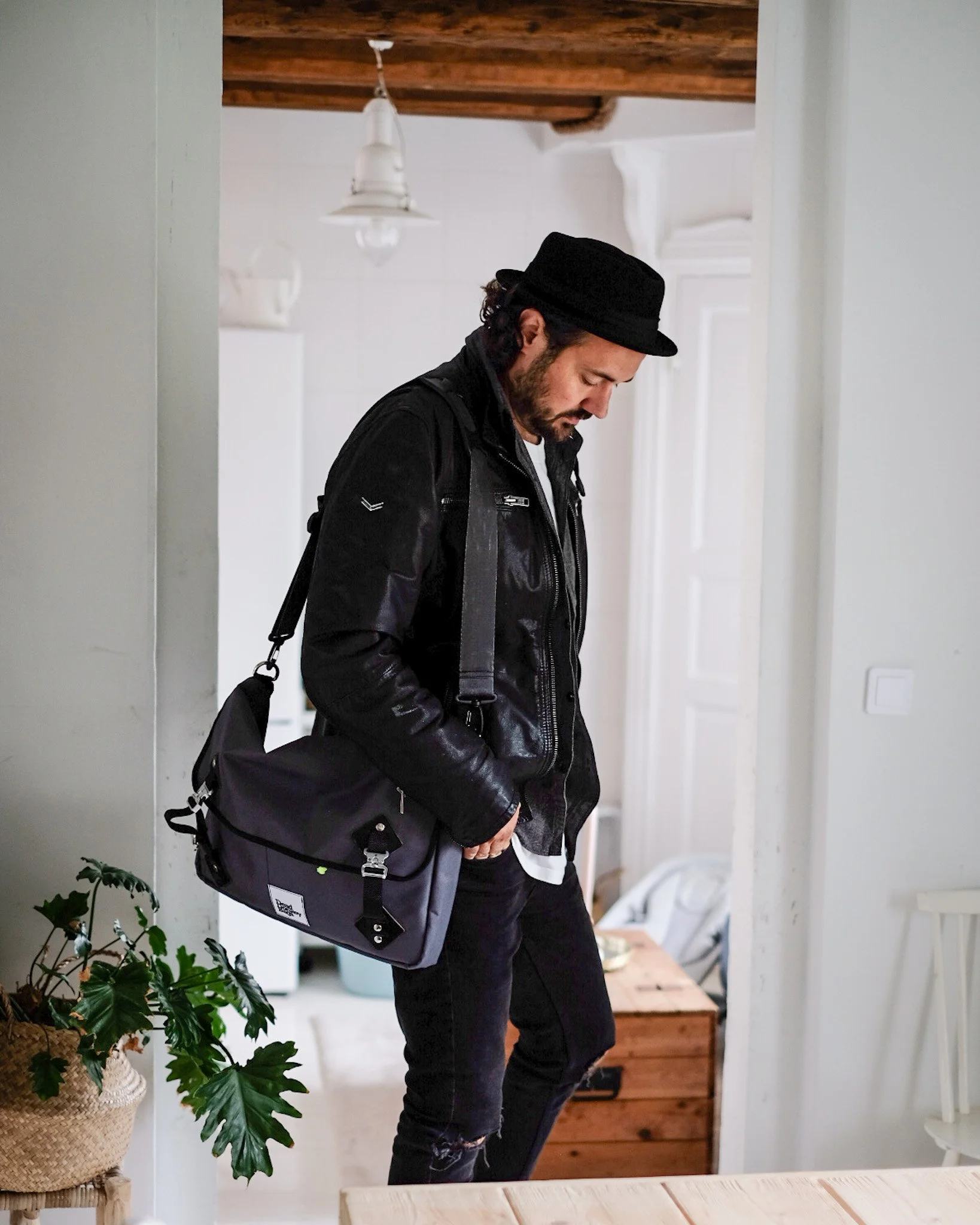 A man with dark hair and beard, dressed in a black leather jacket, black hat, and ripped jeans, standing indoors with his hands in his pockets, carrying a gray shoulder bag.