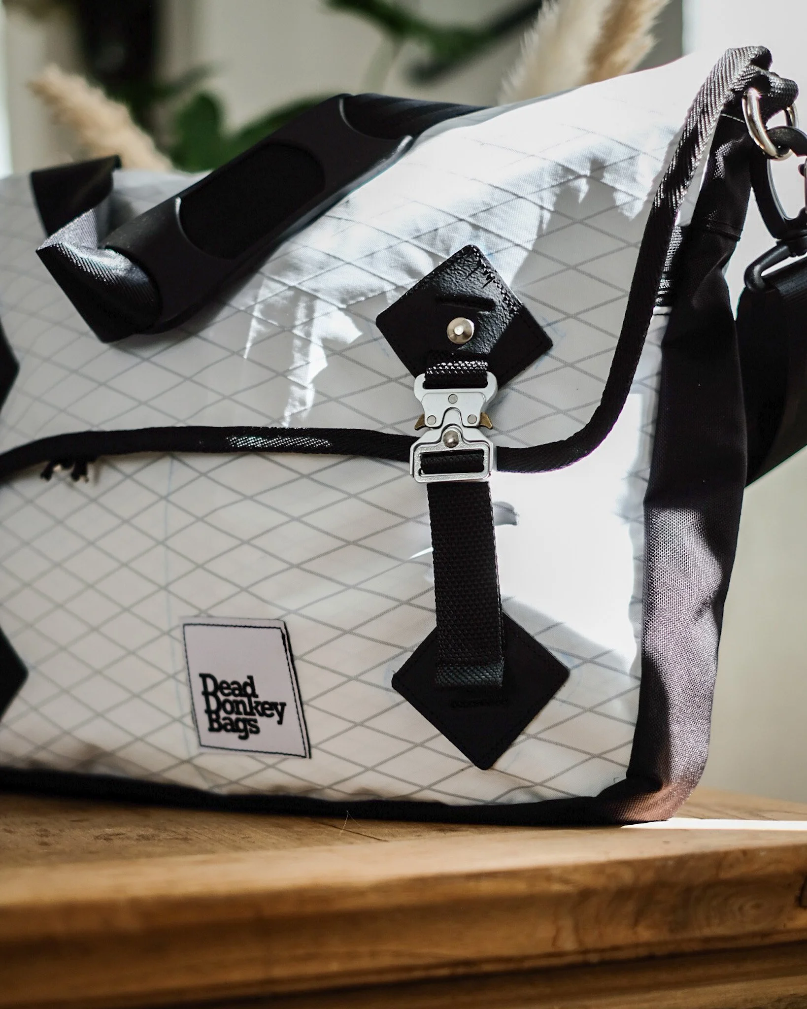 Close-up of a white Dead Monkey Bags backpack with black straps and accents, featuring a diamond grid pattern and metal hardware, placed on a wooden surface.