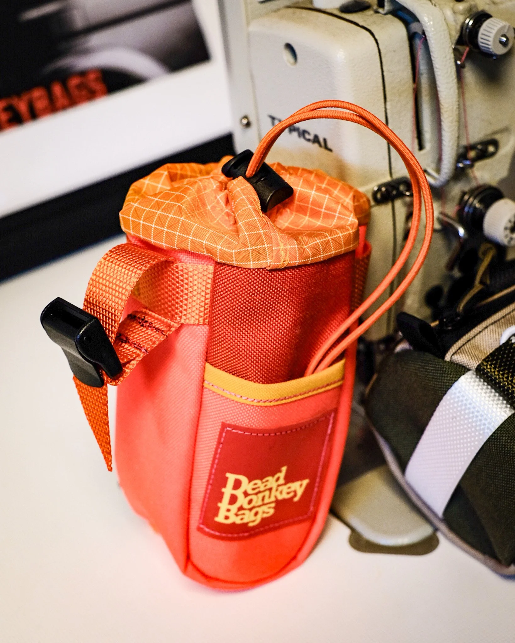An orange and red bag labeled "Dead Monkey Bags" hanging on a hook near a grey machine with knobs and a handle.