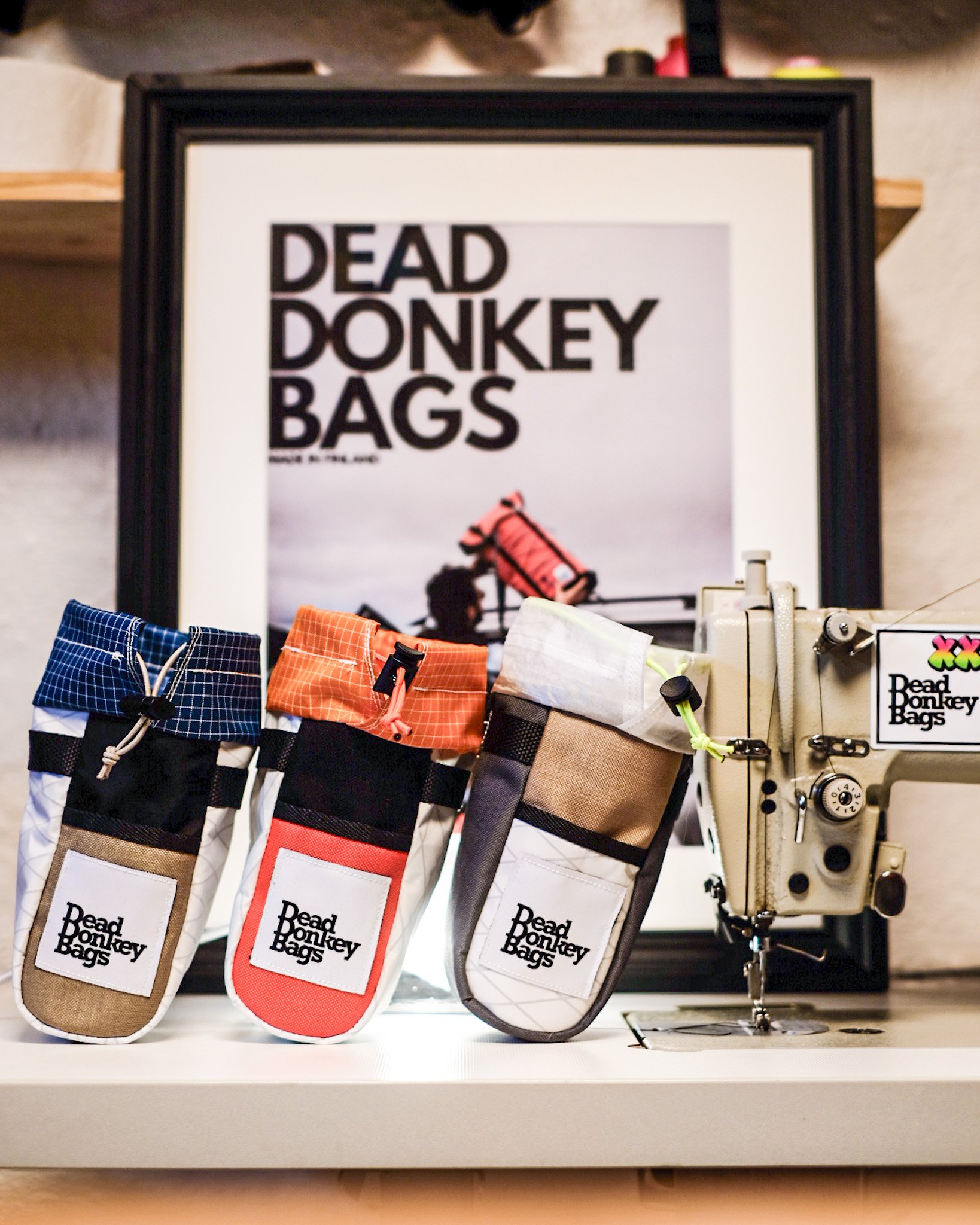 Display of colorful branded bags labeled 'Dead Donkey Bags' in front of a framed poster with the same phrase, and a sewing machine to the right.