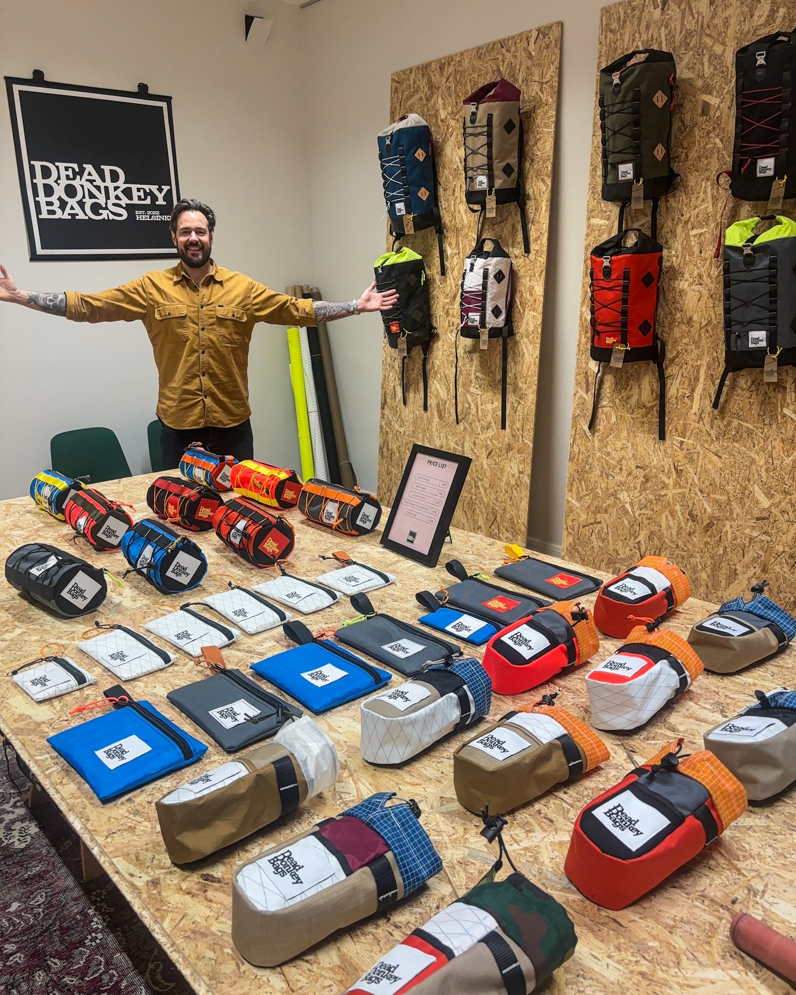 A man standing behind a table displaying various small bags and pouches in different colors and styles, with backpacks hanging on the wall behind him at a store showcasing Reed Boney Bags.