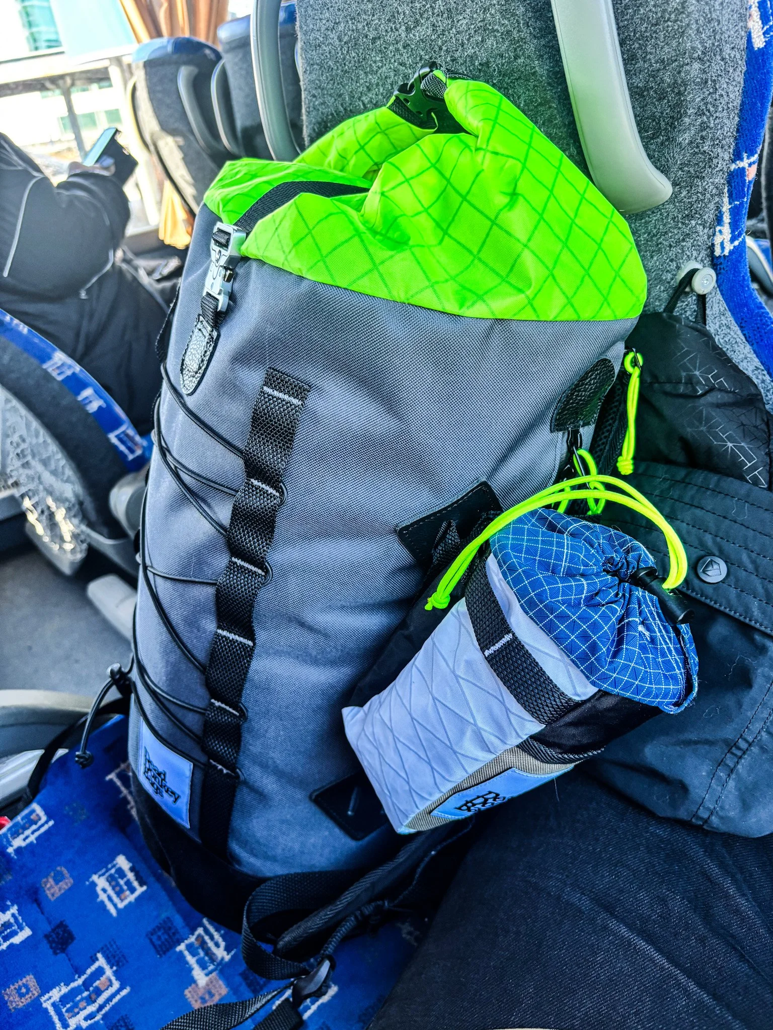 A gray backpack with a green drawstring top, secured with black straps, placed on a bus seat. Next to it is a smaller gray and black bag with a blue drawstring and yellow cord. A person in black is sitting nearby, looking at their phone.