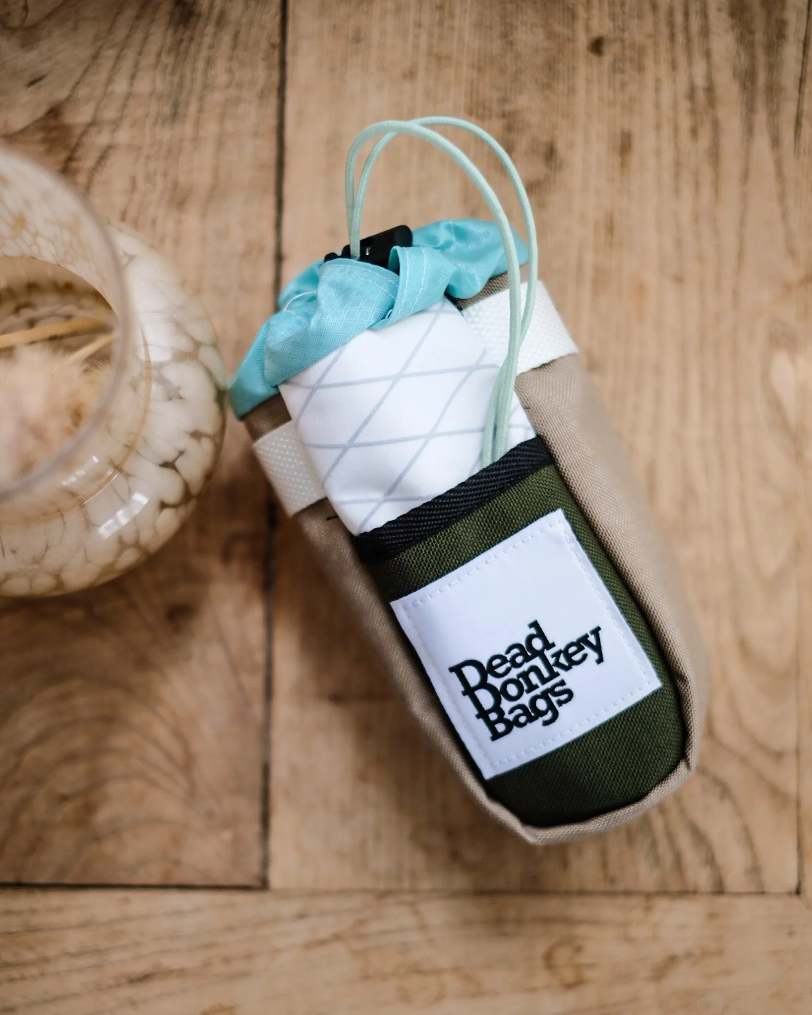 A drink insulator with the label 'Dead Pony Bags' on it, containing a white beverage with ice and a light blue straw, placed on a wooden surface.