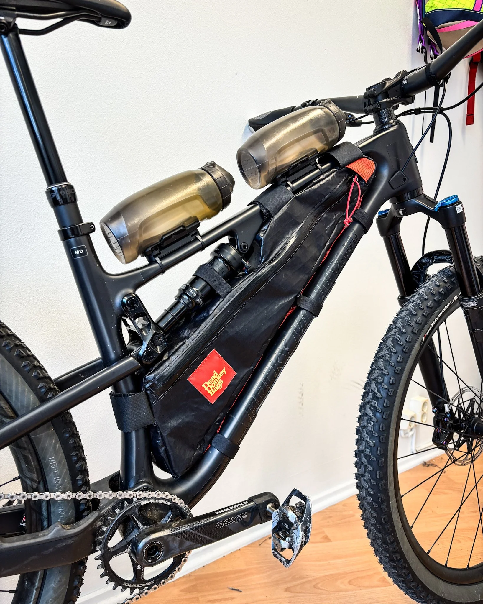 Close-up of a black mountain bike with two water bottles mounted on the top tube, and a large black battery pack attached to the frame, near the bottom bracket. The bike has knobby tires, a suspension fork, and a visible pedal and chainring.