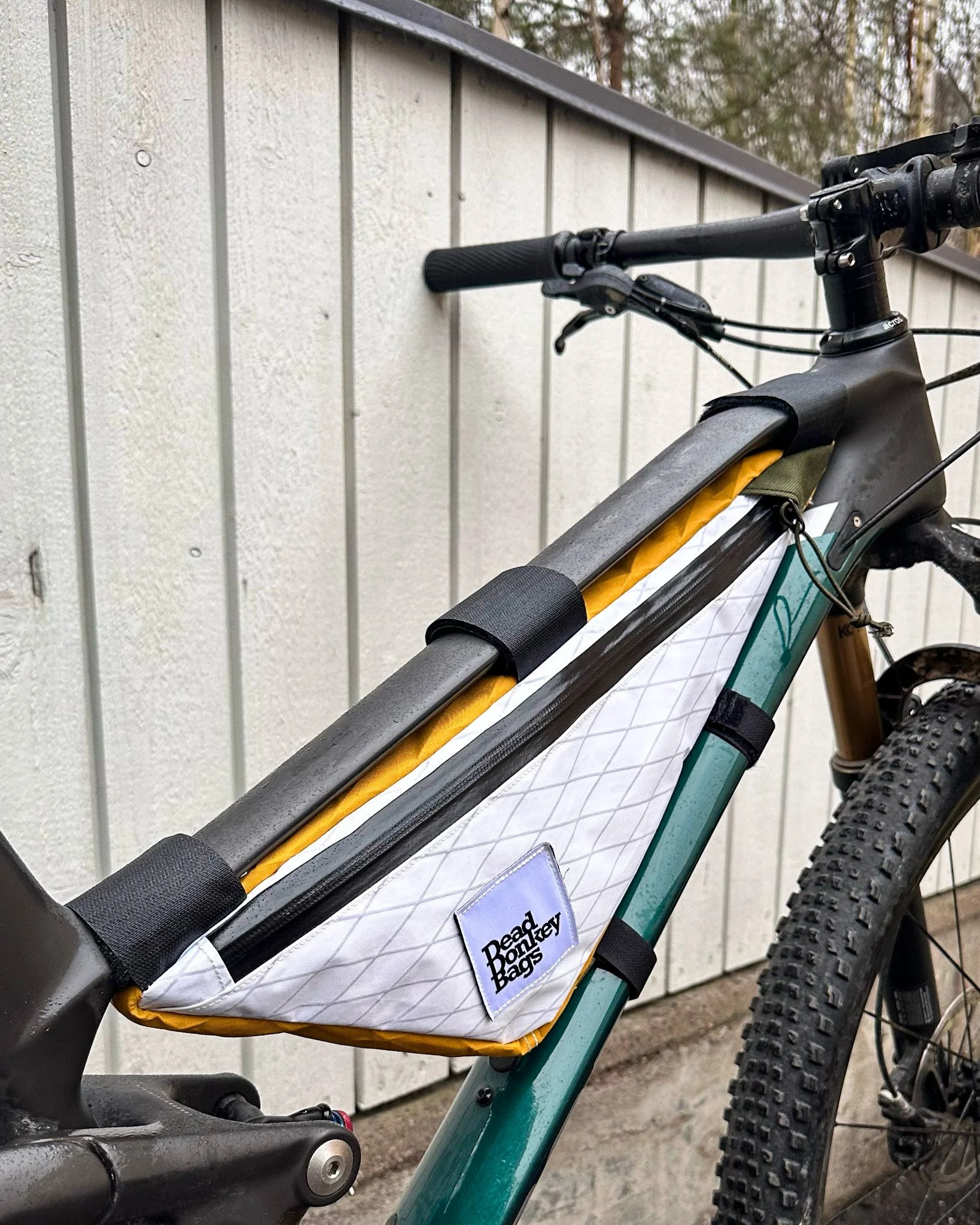 A bicycle with a waterproof bag mounted on the top tube, labeled 'Dead Quarter Bags'. The bag is mainly white with some yellow and black trim, attached with black straps, and positioned against a beige fence with vertical siding.