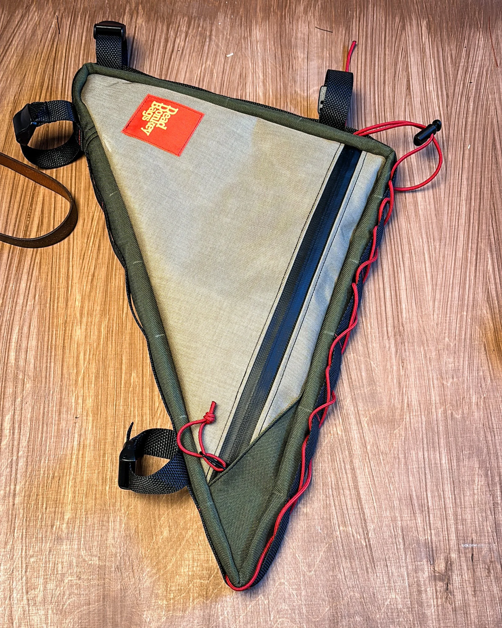 A triathlon bike saddle bag with a beige and green color scheme, featuring a red logo label, a zipper in the middle, and red elastic cords on the edges, resting on a wooden surface.