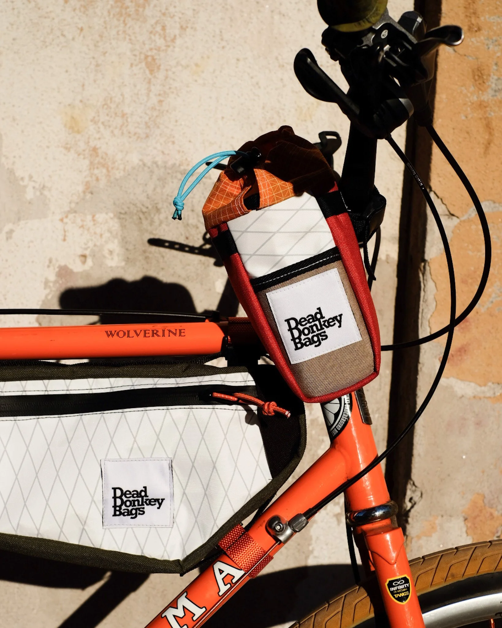 Close-up of an orange bicycle with Dead Donkey Bags attached to the frame, one small bag on the handlebar and one large bag on the top tube, both labeled Dead Donkey Bags.