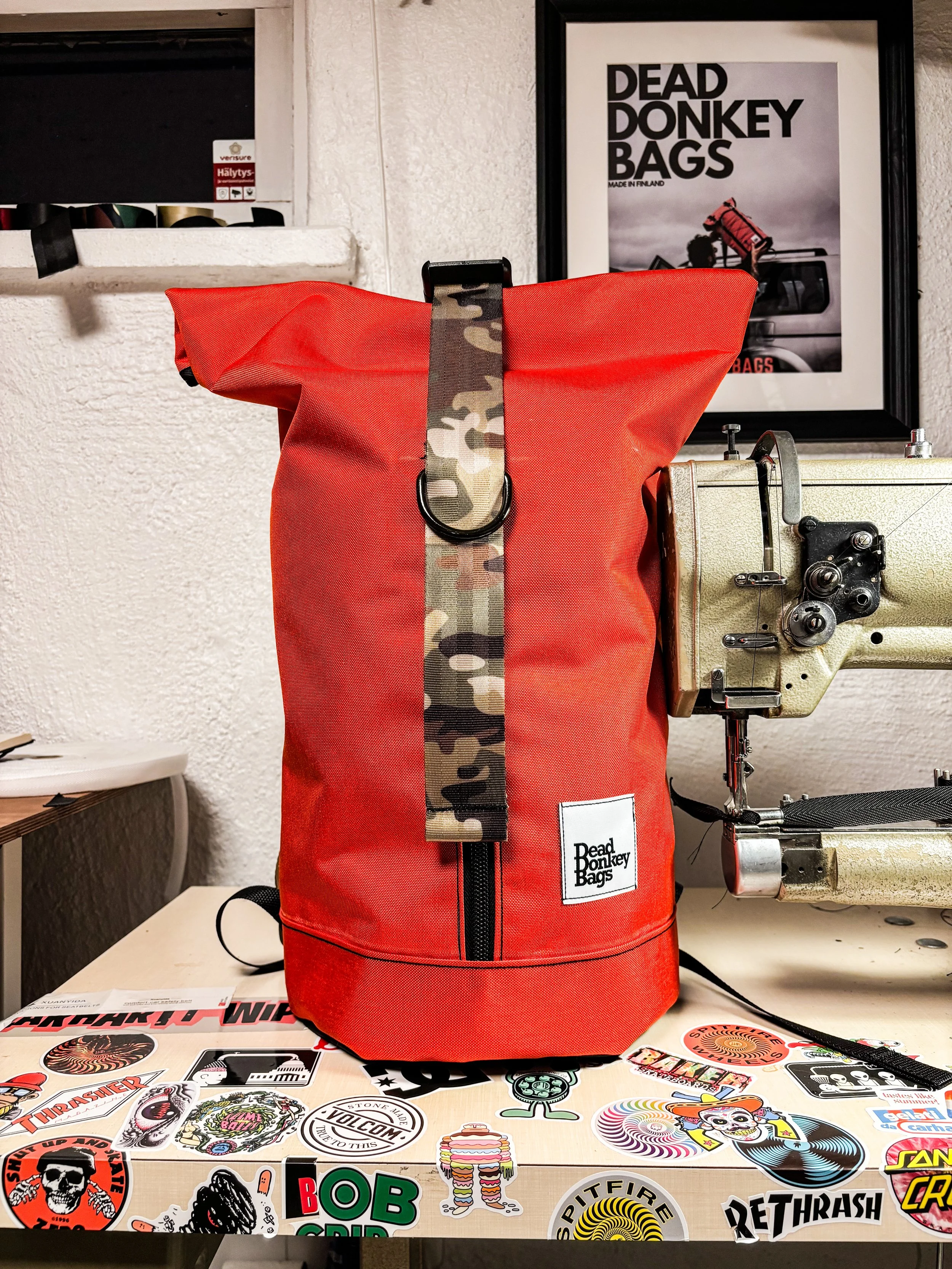 A red Dead Donkey Bags backpack with a floral strap and a black D ring, sitting on a desk with various stickers, next to a sewing machine, with a framed Dead Donkey Bags poster on the wall behind.