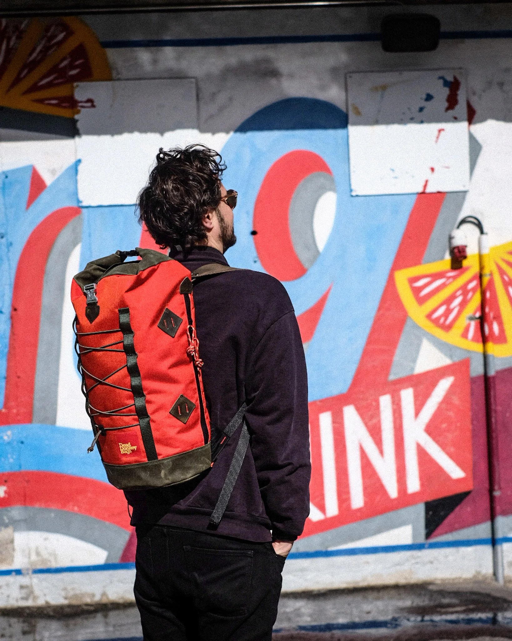 A man with dark curly hair, wearing sunglasses, a black jacket, and black pants, standing outdoors with his hands in his pockets, looking at a colorful graffiti mural on a wall. The mural features the words 'No Stink' in bold letters with bright red,