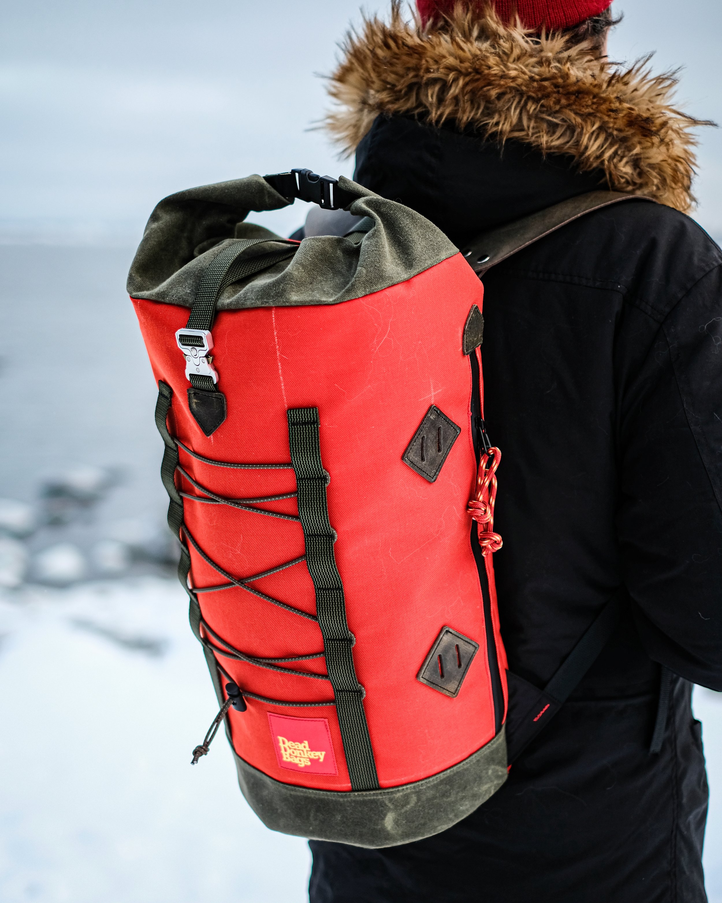 Person wearing a black jacket with a fur-lined hood carrying a red and gray dry bag on their back outdoors in snowy terrain.