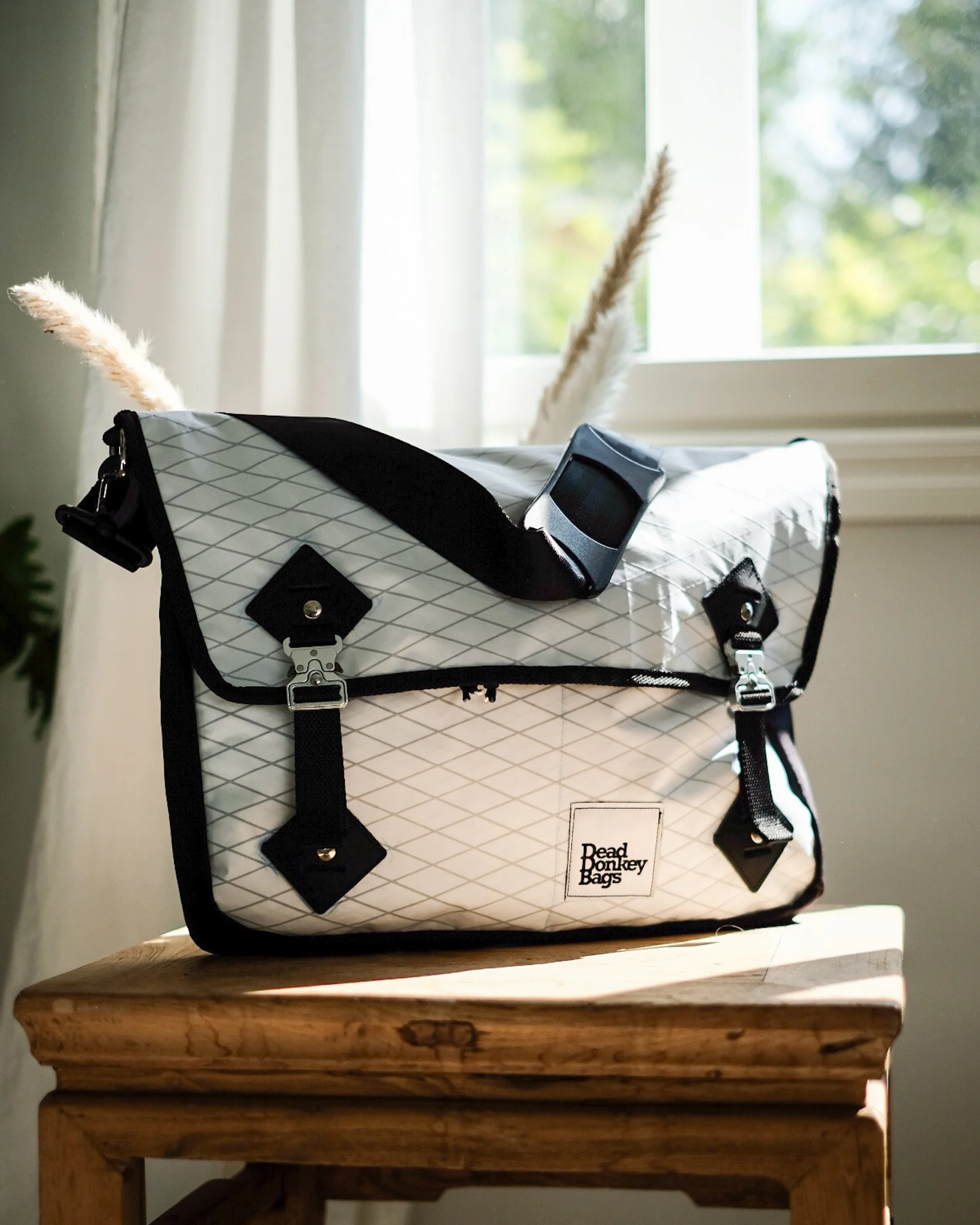 A white and black Dead Bonkey Bags bicycle messenger bag on a wooden surface near a window with sunlight and green trees outside.