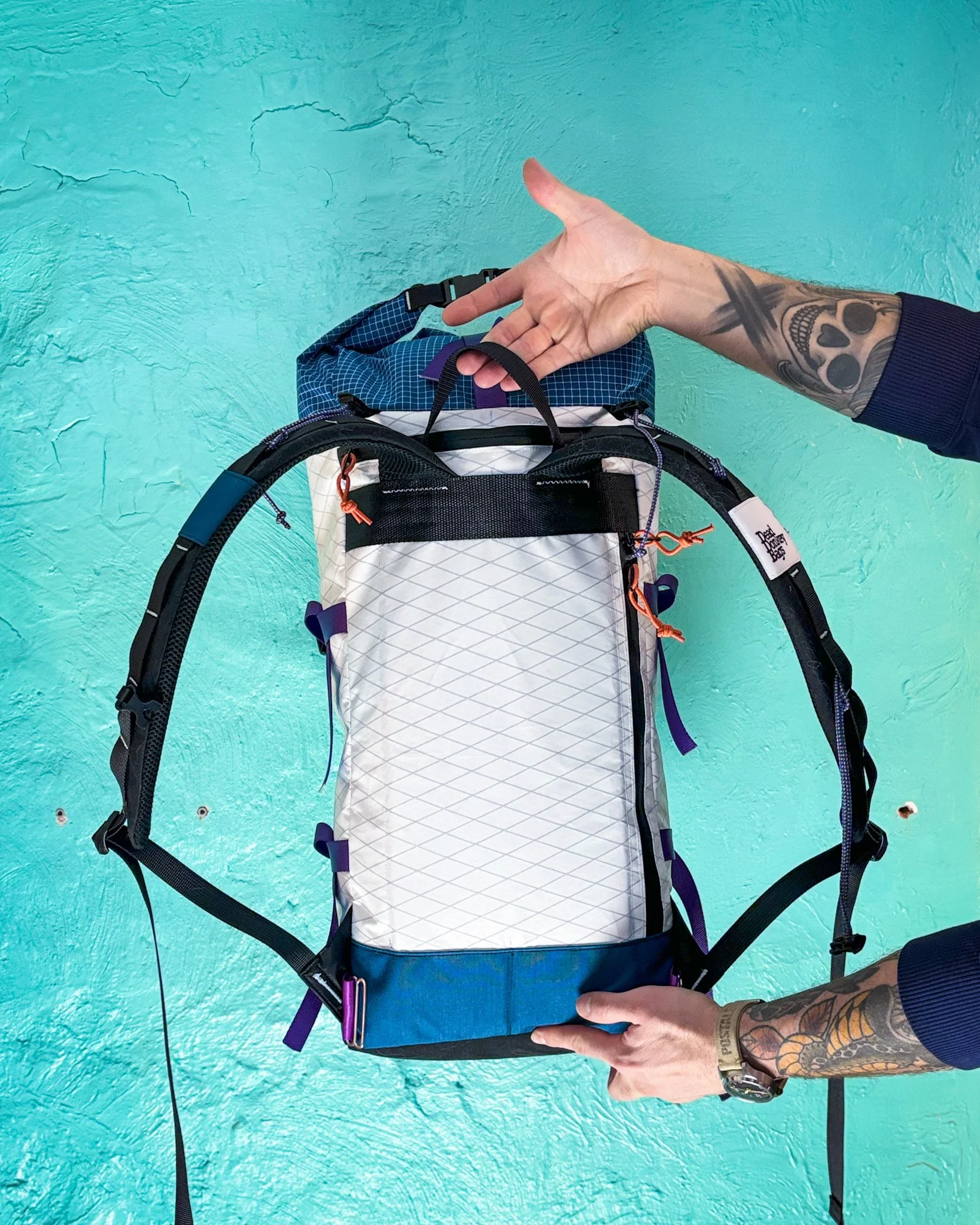 Person holding a white and blue hiking backpack against a teal textured wall, showing colorful straps and zippers.