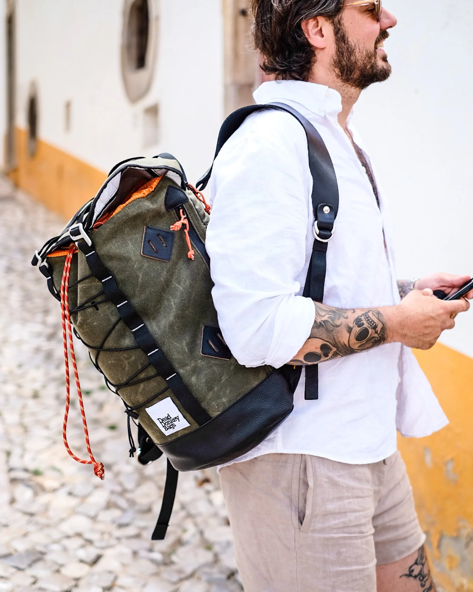 A man with tattoos on his arm wearing a white shirt and khaki shorts, carrying a green and black backpack, and looking at his phone.