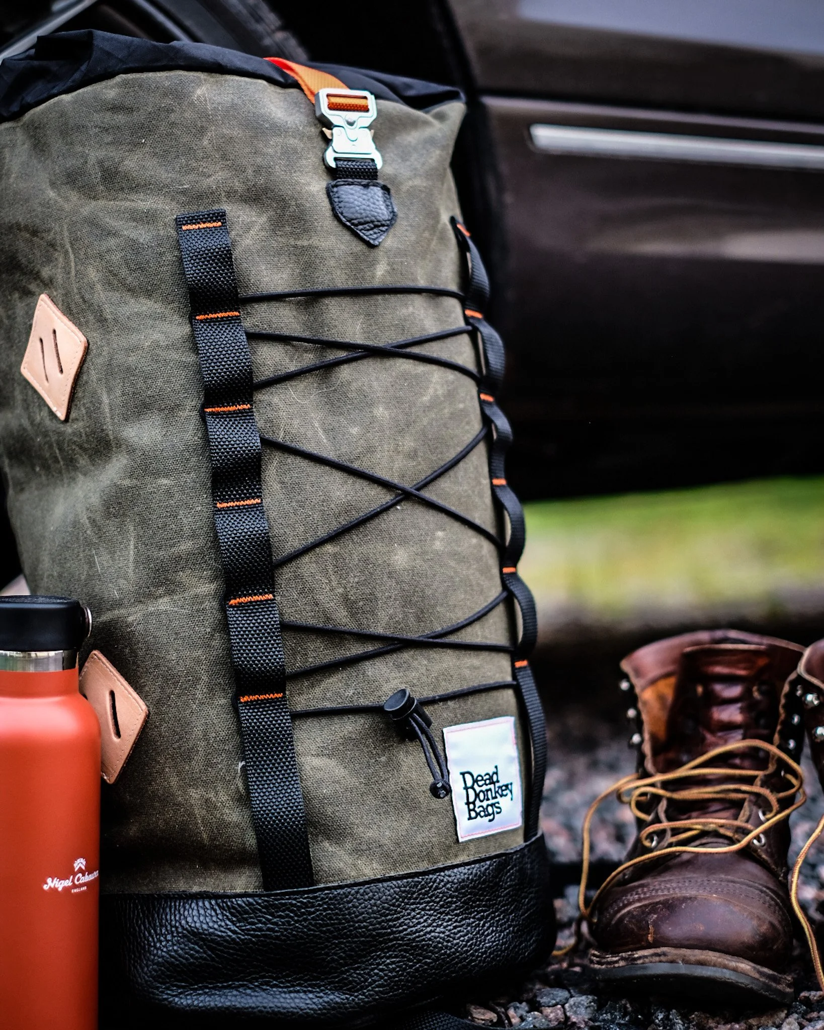 A greenish-brown backpack with black straps and a label that reads 'Dead Donkey Bags,' a brown rugged hiking boot with yellow laces, and a copper-colored travel mug with a black lid, placed outdoors on a gravel surface.