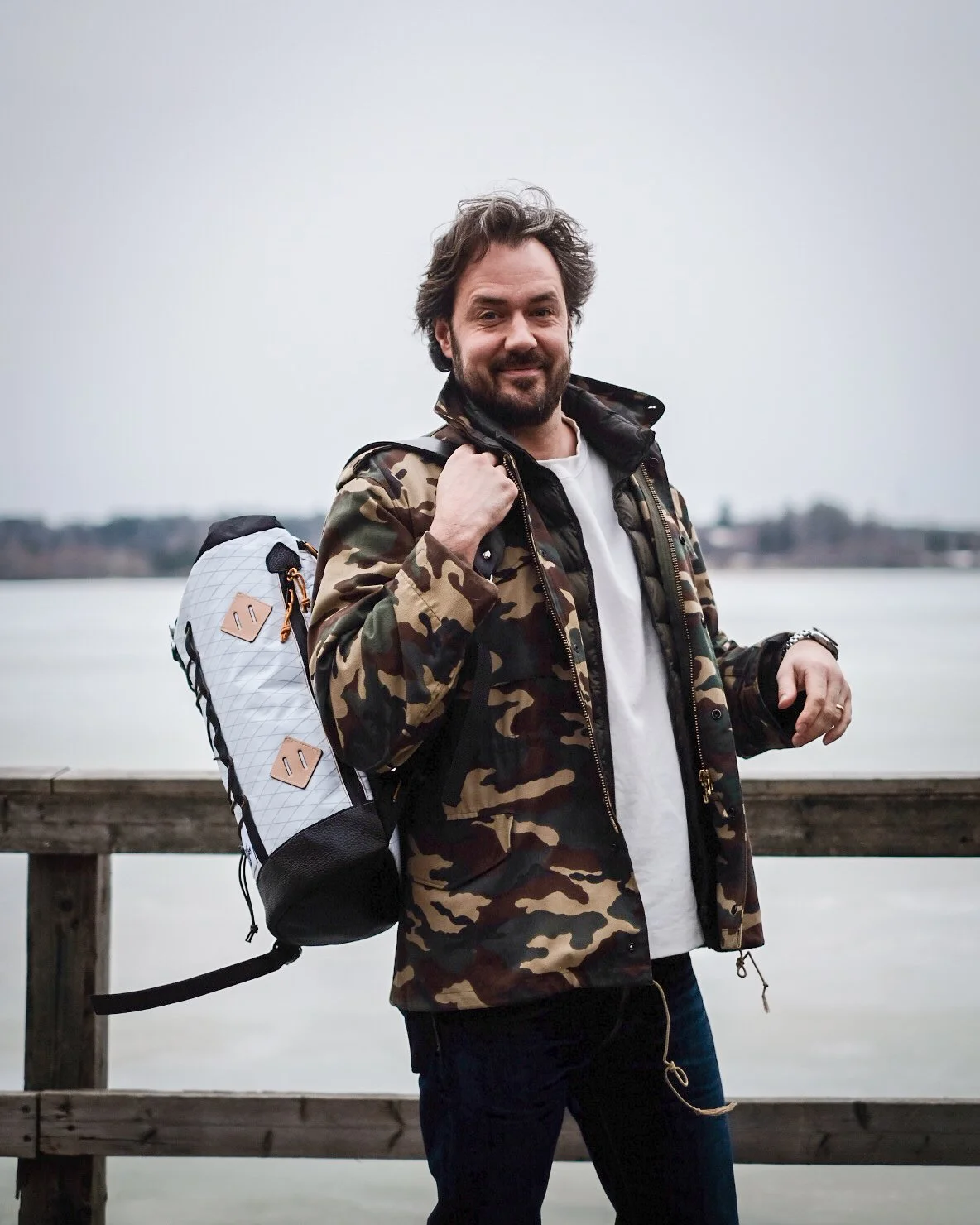 A man with brown hair and a beard stands on a wooden dock, carrying a backpack, wearing a camouflage jacket over a white shirt, near a body of water on a cloudy day.