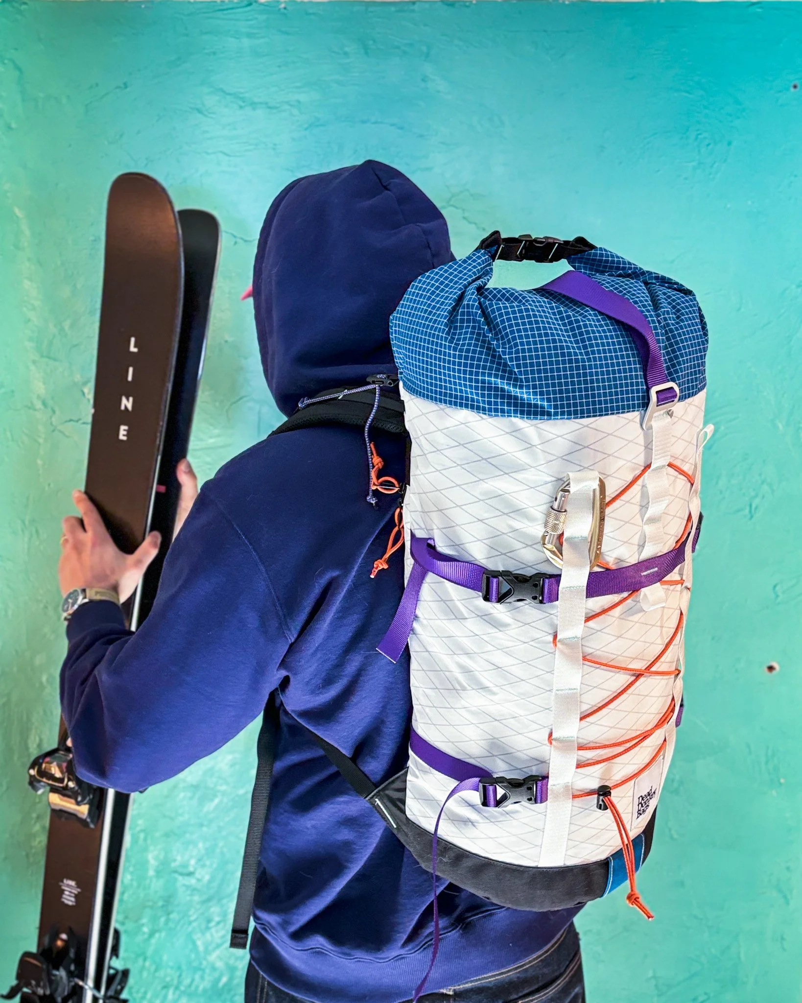 Person dressed in a navy hoodie holding skis, standing against a turquoise wall, with a large multicolored backpack on their back.
