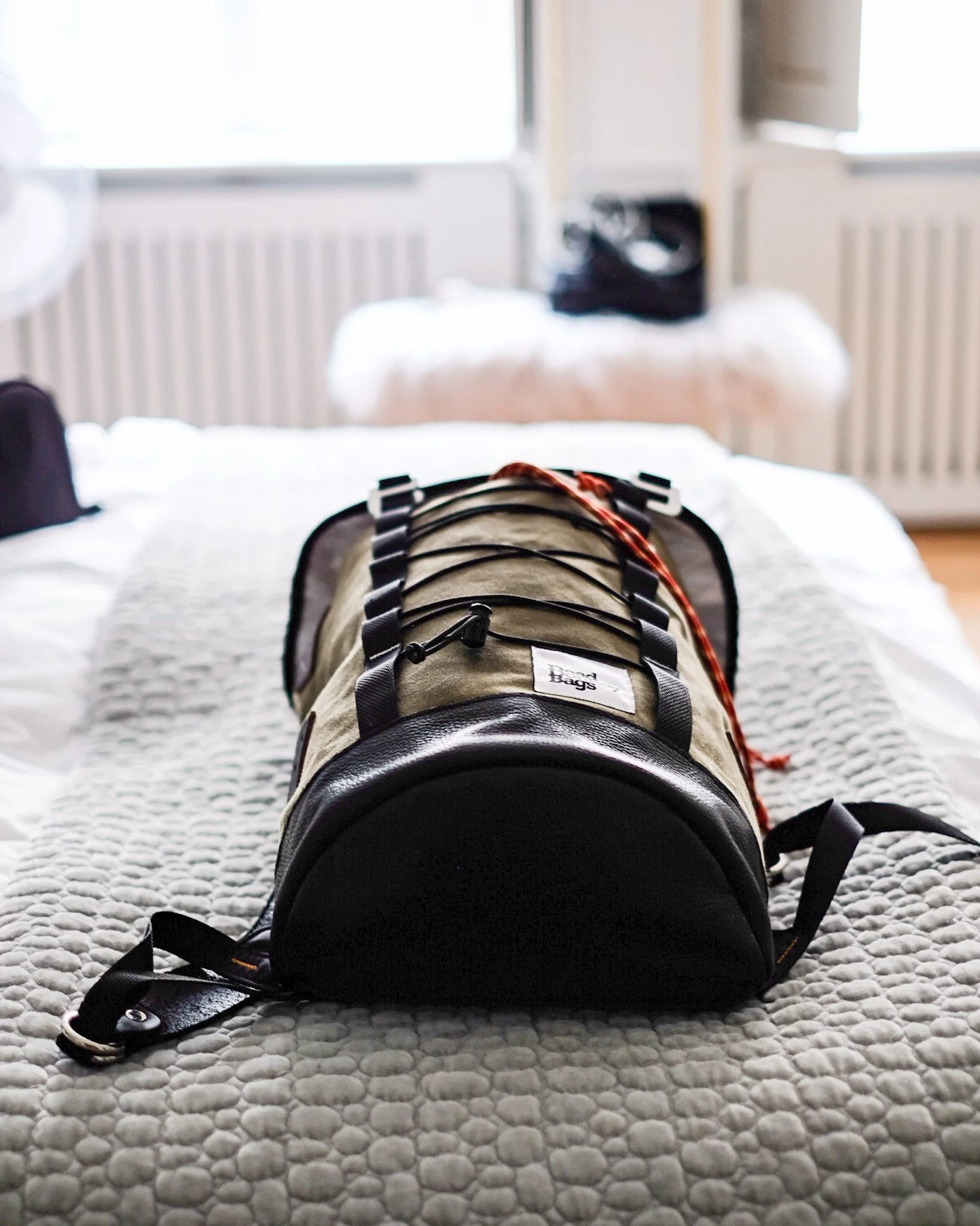 A duffel bag with black straps and leather ends on a bed with a textured gray quilt, in a bright room with large windows.