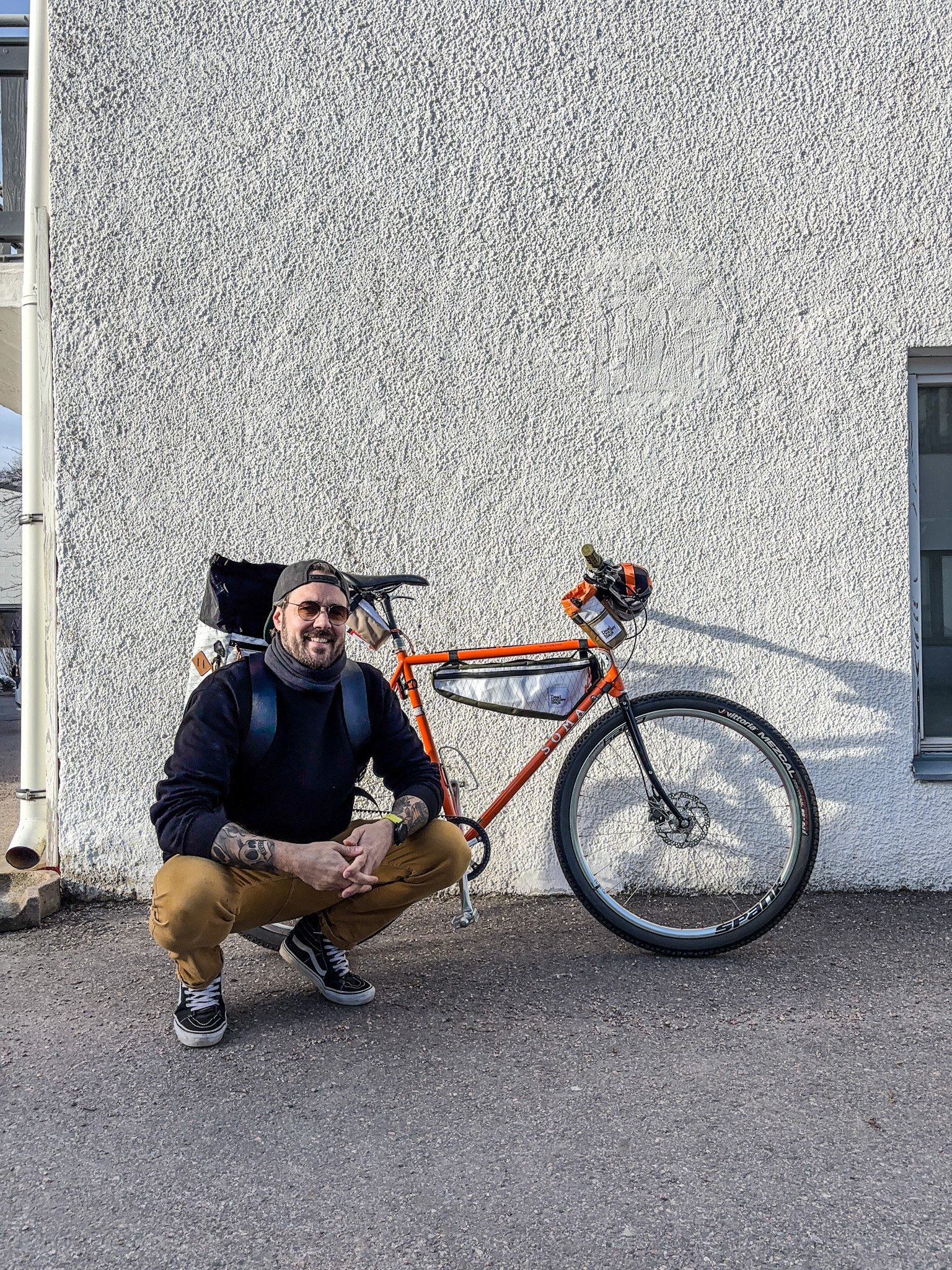 A man with tattoos, wearing sunglasses, kneeling beside an orange and black bicycle against a textured white wall.
