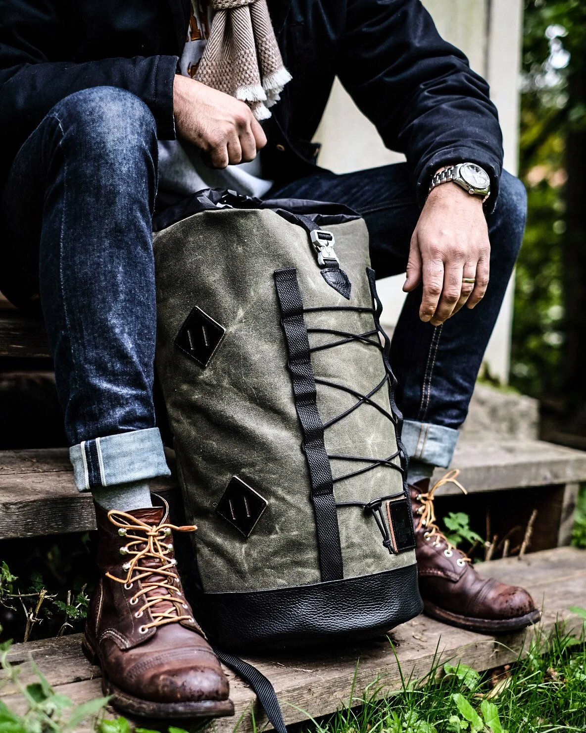 Person sitting outdoors on a wooden bench, wearing jeans, a black jacket, and boots, with a large olive green backpack on the ground in front of them.