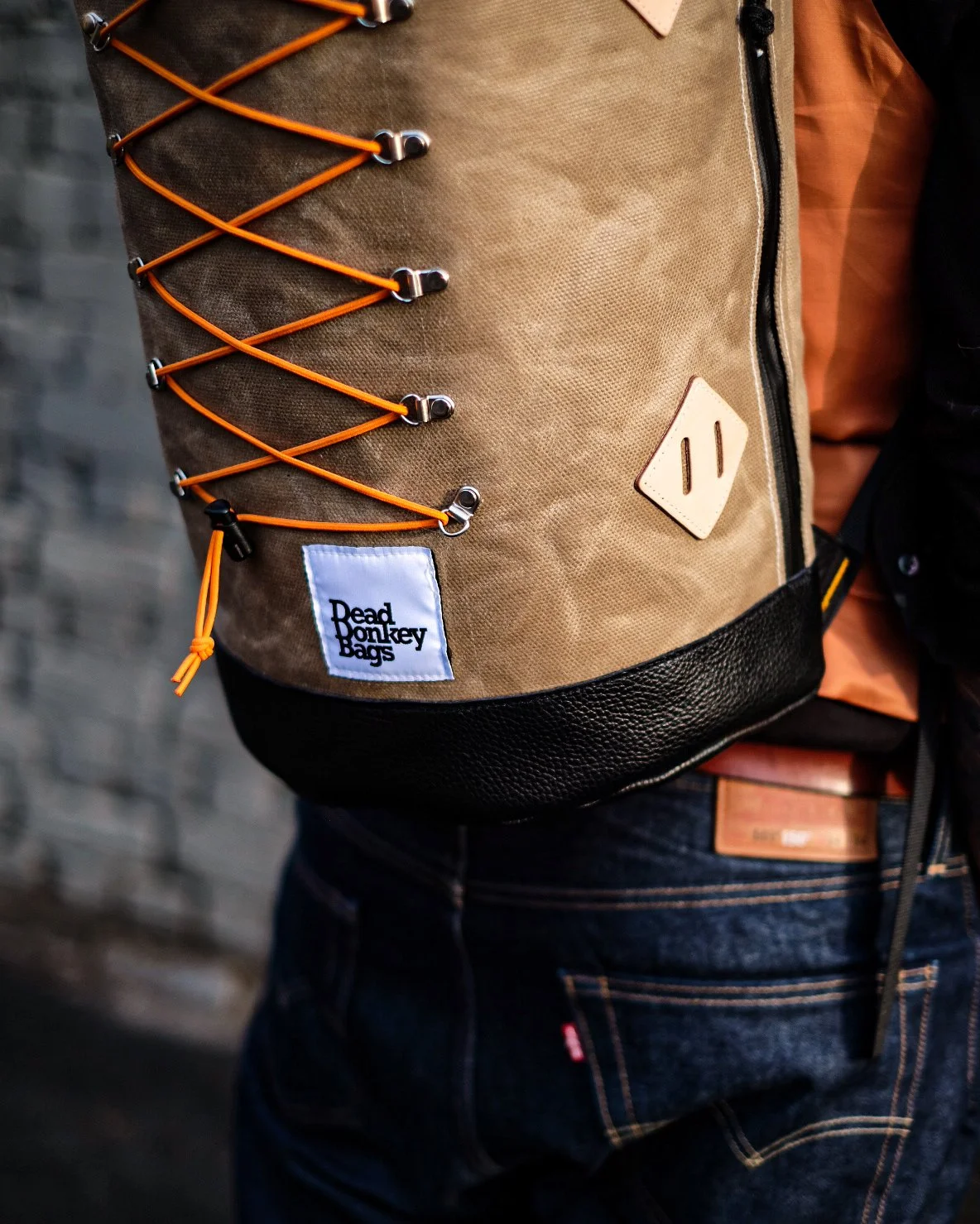Person wearing a tan backpack with orange laces, a logo patch that reads 'Read Monkey Bags,' and a beige triangular patch, in front of a dark brick wall.