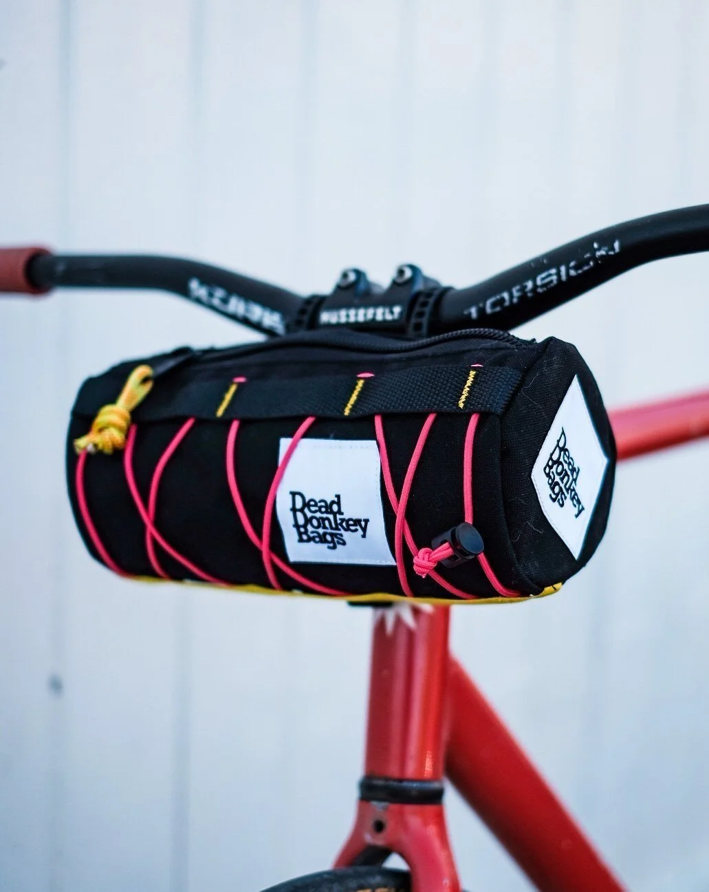 A black bicycle handlebar with a black bag attached to the front, labeled 'Dead Monkey Bags', mounted on a red bicycle frame.