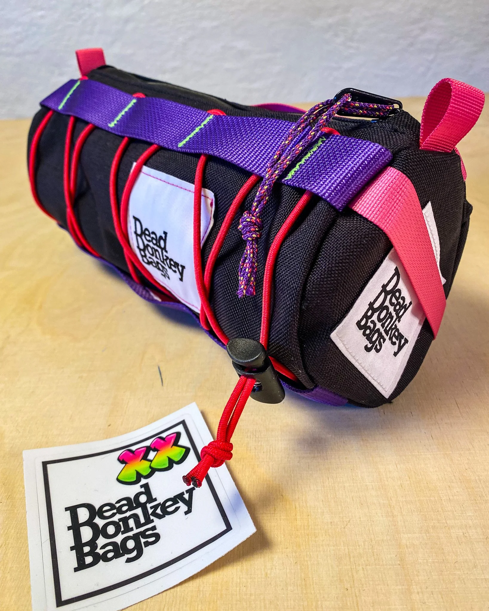 A small black tool roll with red elastic bands, pink and purple straps, and a white label with the logo 'Dead Monkey Bags'. A colorful tag with the same logo is attached to the roll. The tool roll is placed on a light wooden surface.
