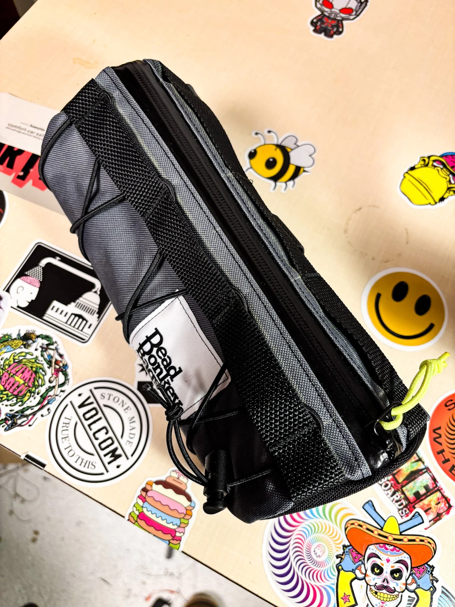 A gray and black utility pouch with zippers, placed on a table decorated with colorful stickers, including a smiley face, bee, and cartoon characters.