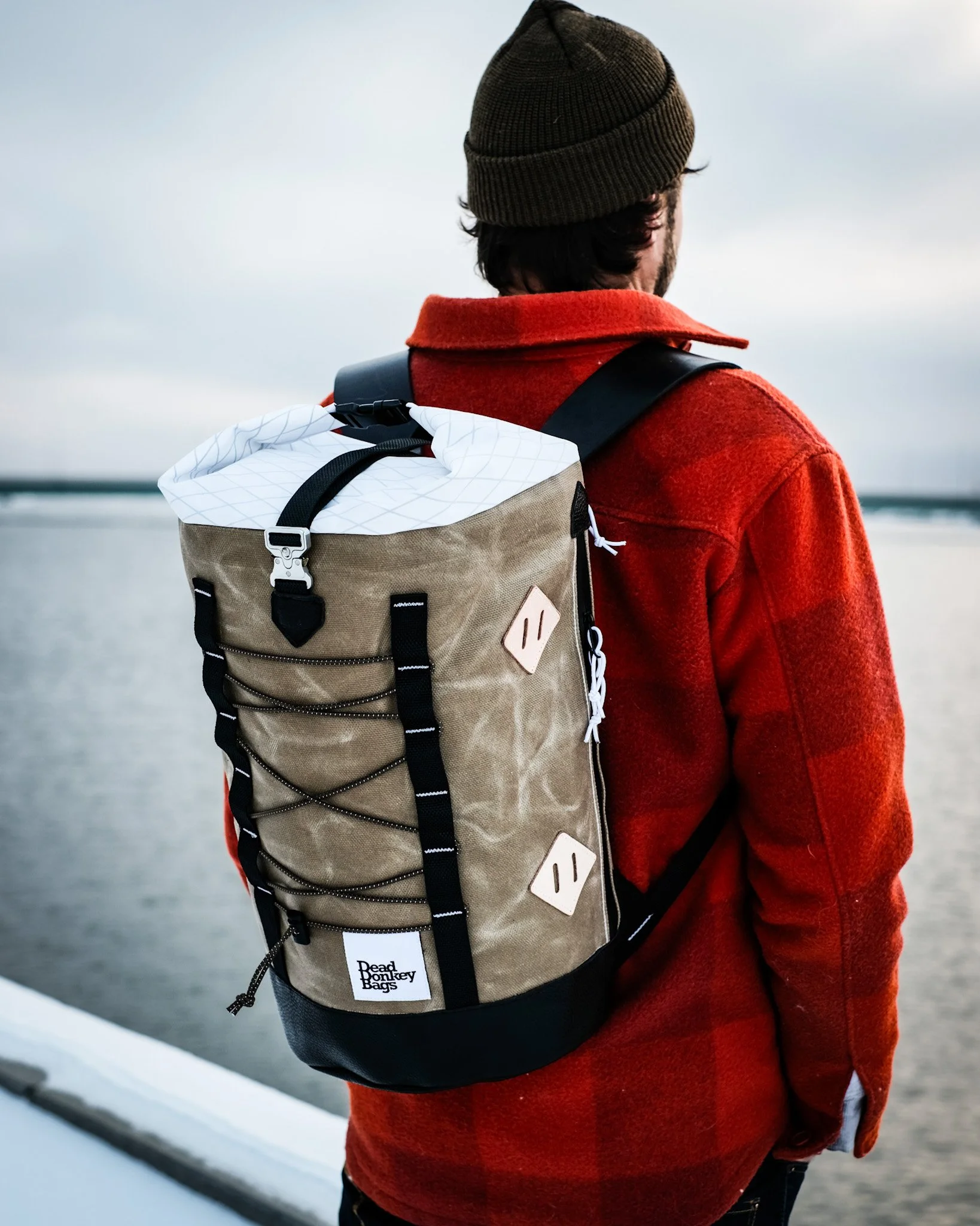 A person wearing a red jacket and brown beanie standing by water, carrying a tan and black backpack with white accents.