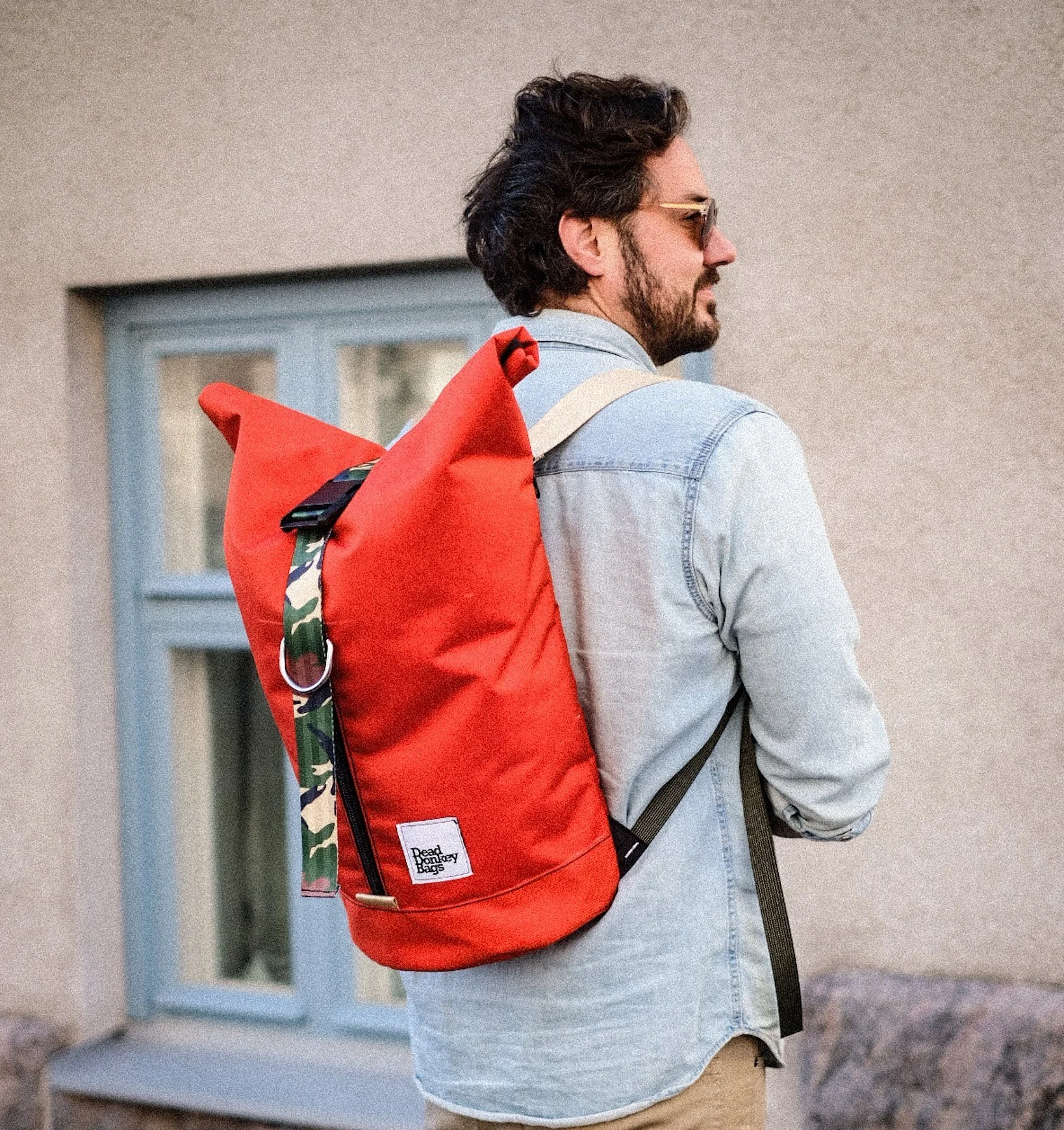 A man with dark hair, beard, and sunglasses, wearing a light denim jacket, carries a red backpack with a camo strap, standing outside near a building with a window.