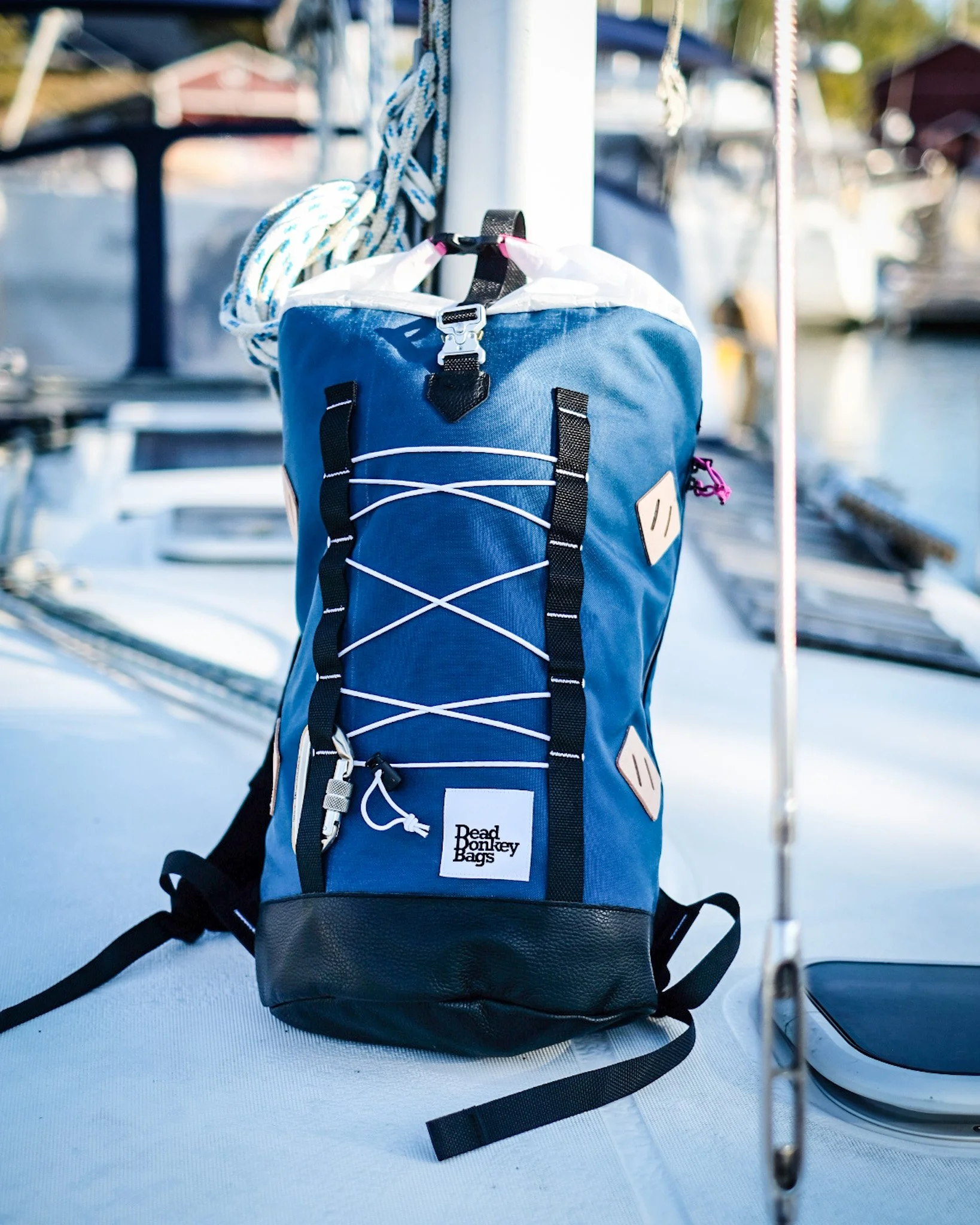 A blue backpack with black straps and a white logo that reads "Dead Donkey Bags" is sitting on a boat, with nautical equipment and other boats visible in the background.