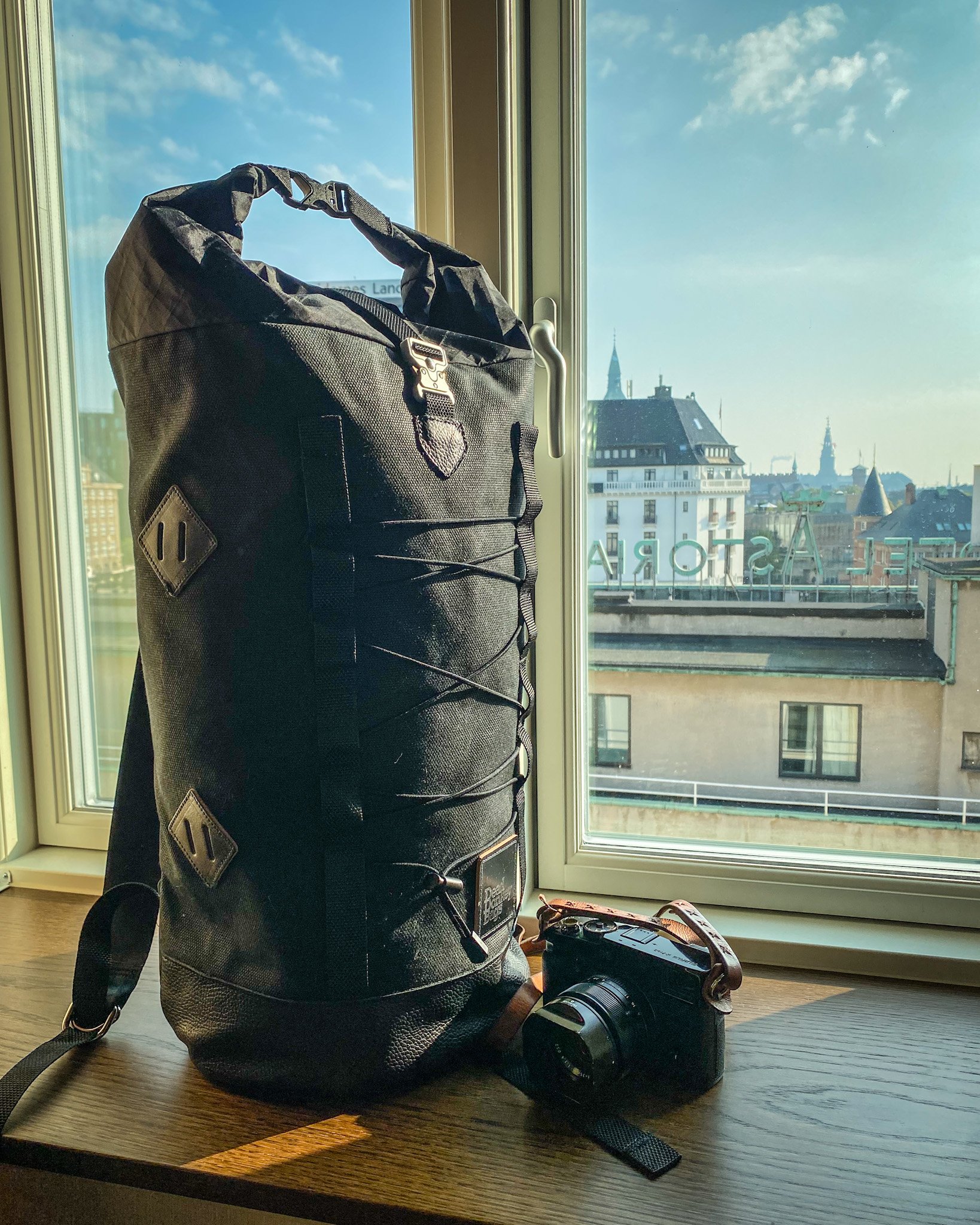 A black backpack and a camera with a strap on a wooden table next to a window overlooking city buildings and spires.