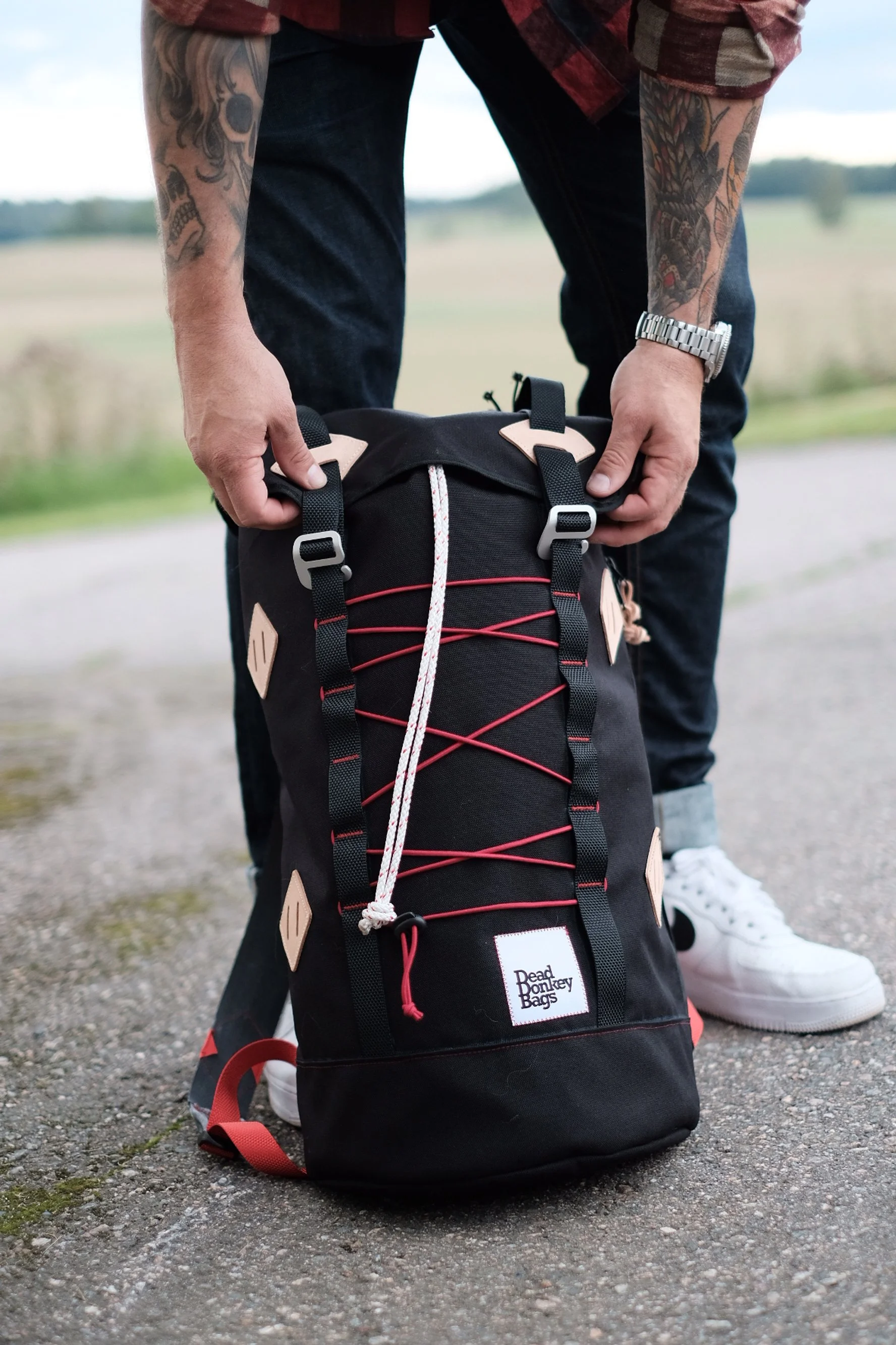 Person adjusting a black Dead Donkey Bags backpack with tan accents and red cord on the front, standing outdoors on a gravel path.