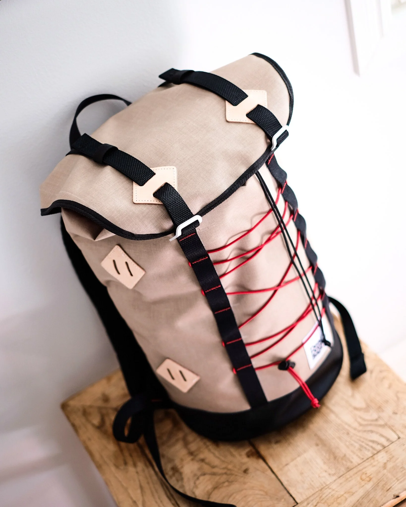 Beige outdoor backpack with red cord laces and black straps, hanging on a wooden surface.