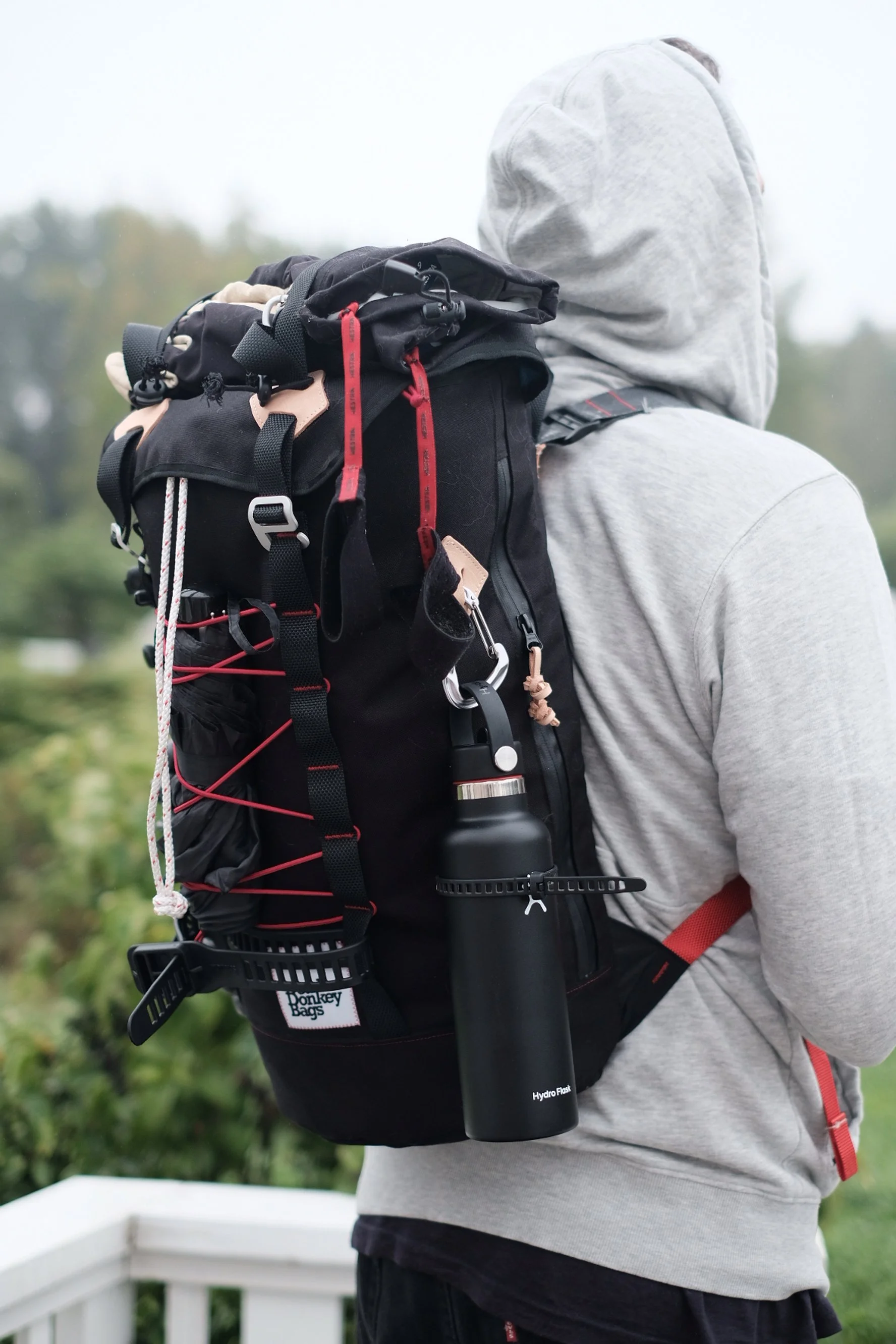Person wearing a gray hoodie and black pants hiking outdoors with a large black backpack that has gear attached, including a black Hydro Flask water bottle.