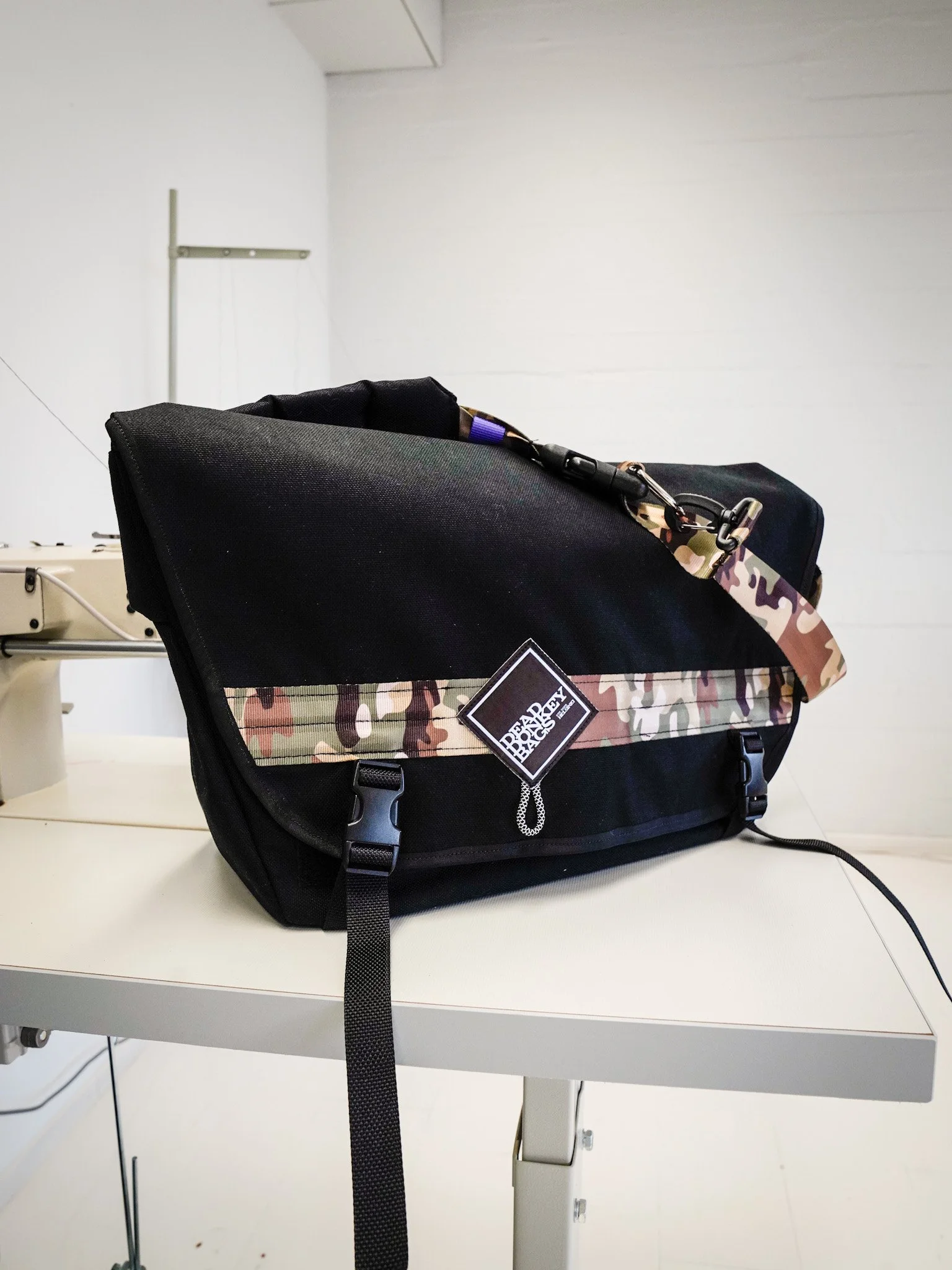 Black bag with a camouflage strap and a Prada label, sitting on a white table in a plain room.